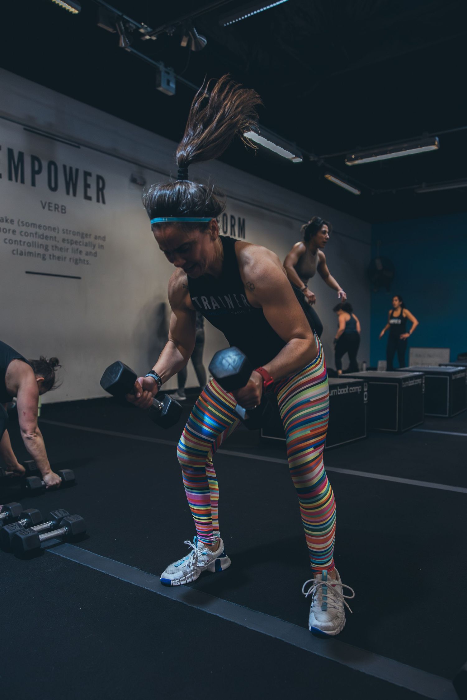 A person in colorful striped leggings works out with dumbbells in a dark fitness studio.