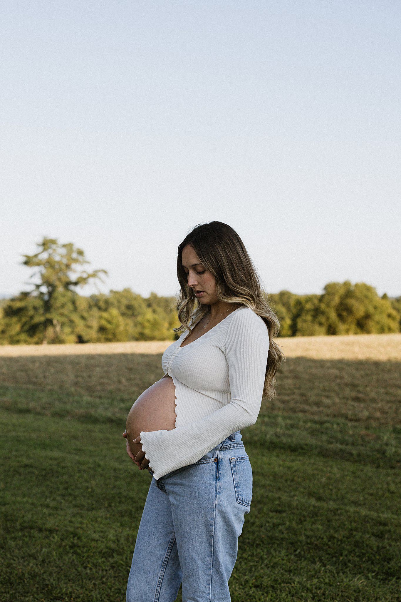 Pregnant woman in white sweater and jeans standing in golden hour light.