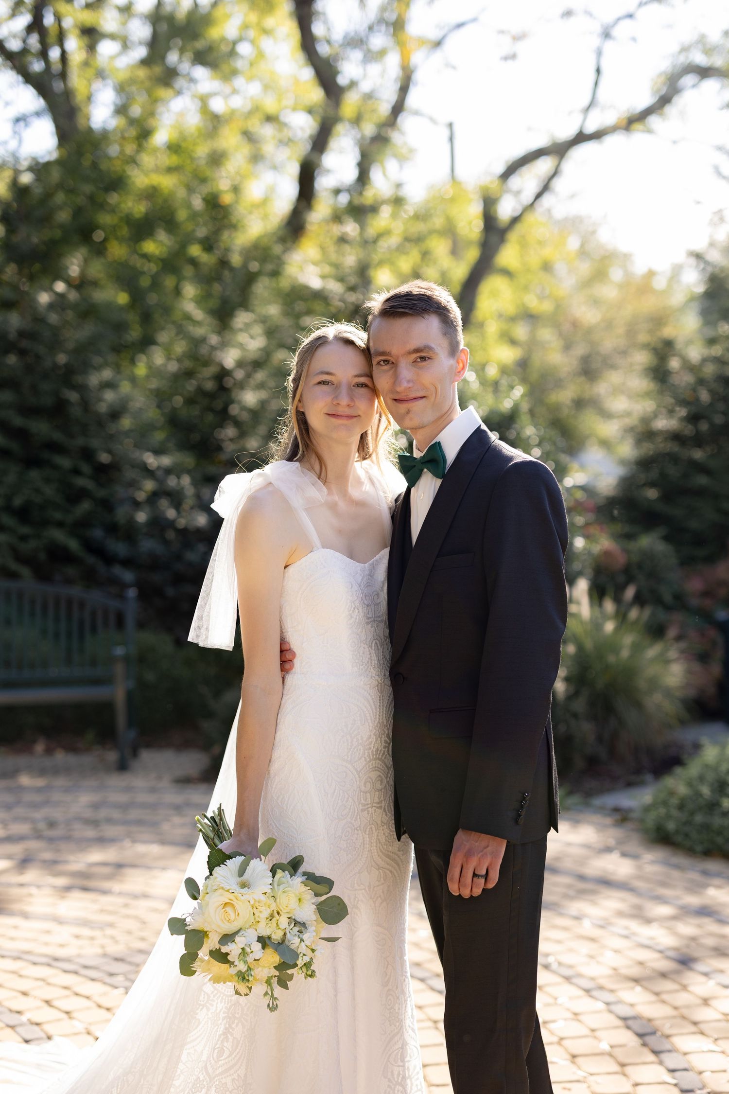 A couple poses together on their wedding day in a garden setting with autumn foliage in the background.