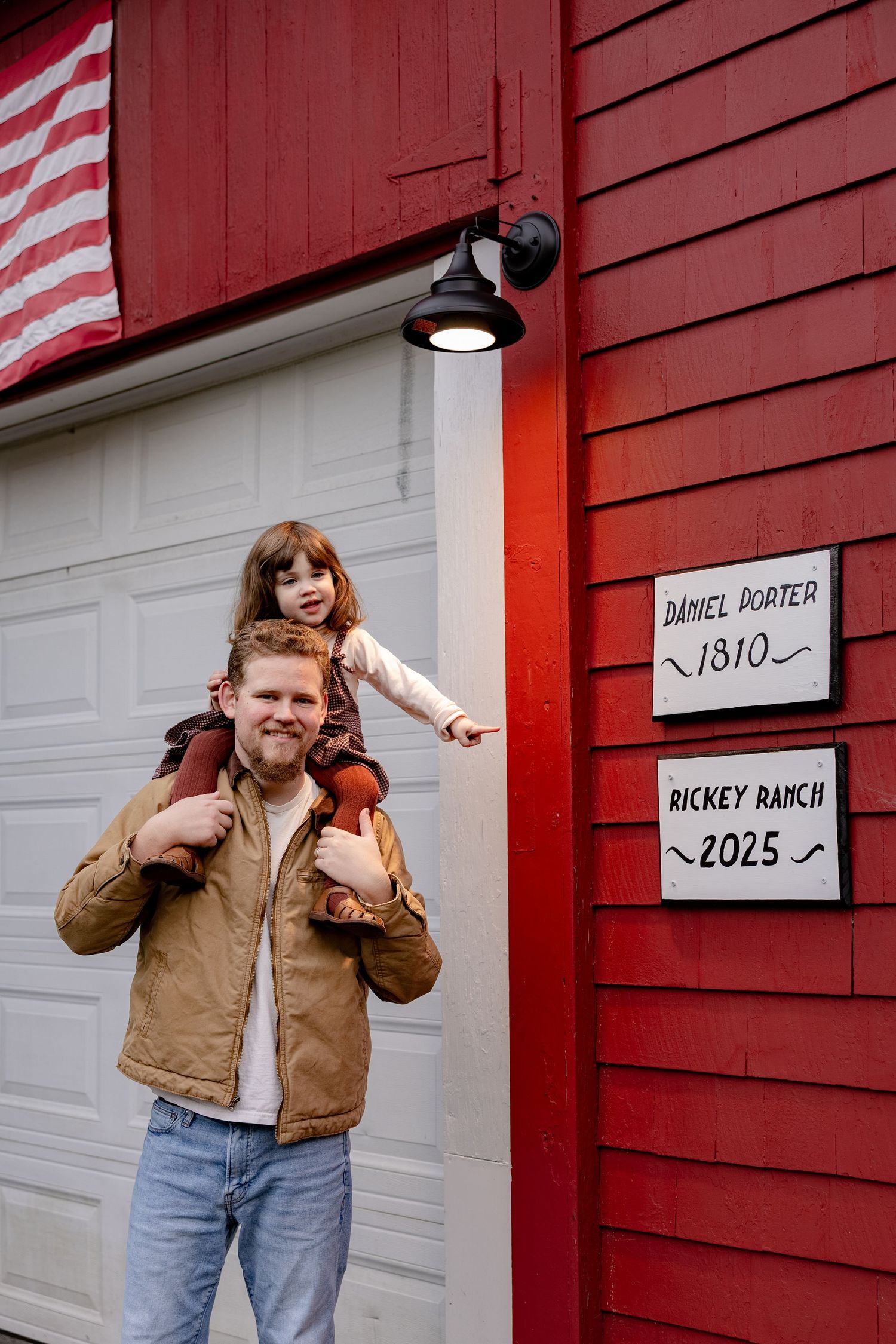 A rustic red barn with a mounted light fixture and two signs showing dates, with an American flag visible in frame.