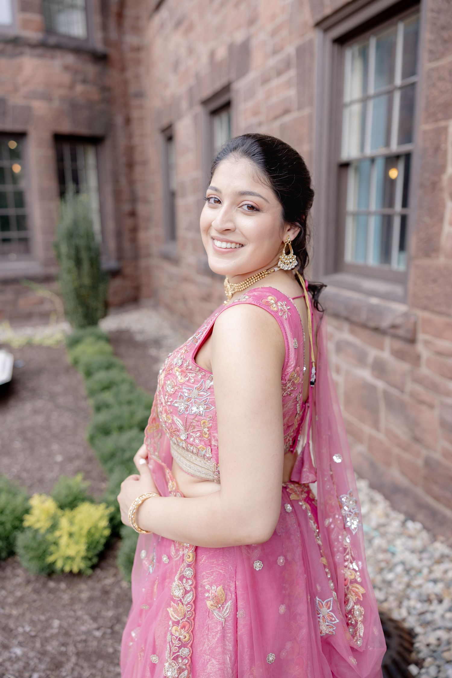 Woman wearing a pink embroidered lehenga poses elegantly against a brick building exterior.
