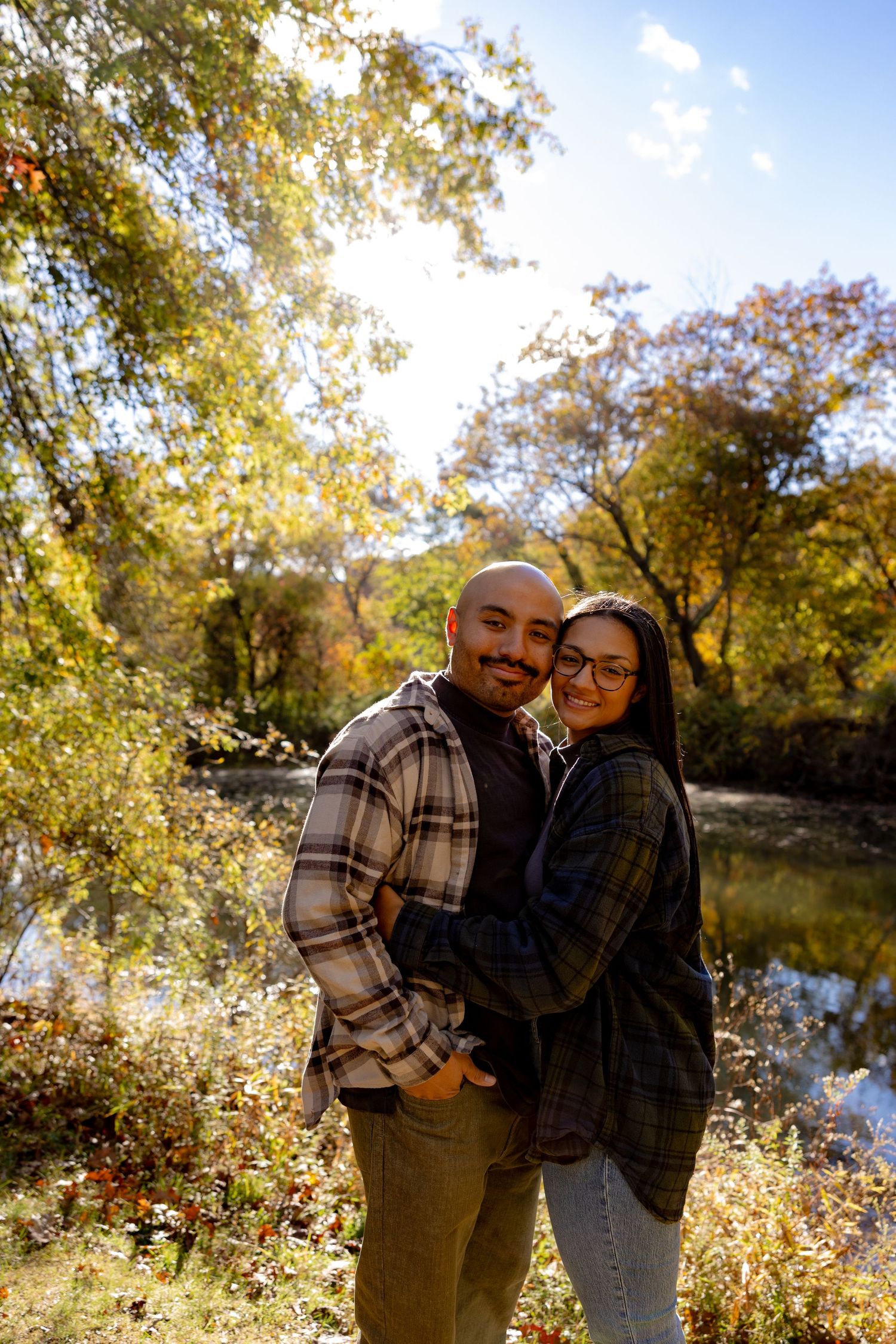 A couple shares an intimate moment together in a sunlit autumn setting by a lake surrounded by golden foliage.