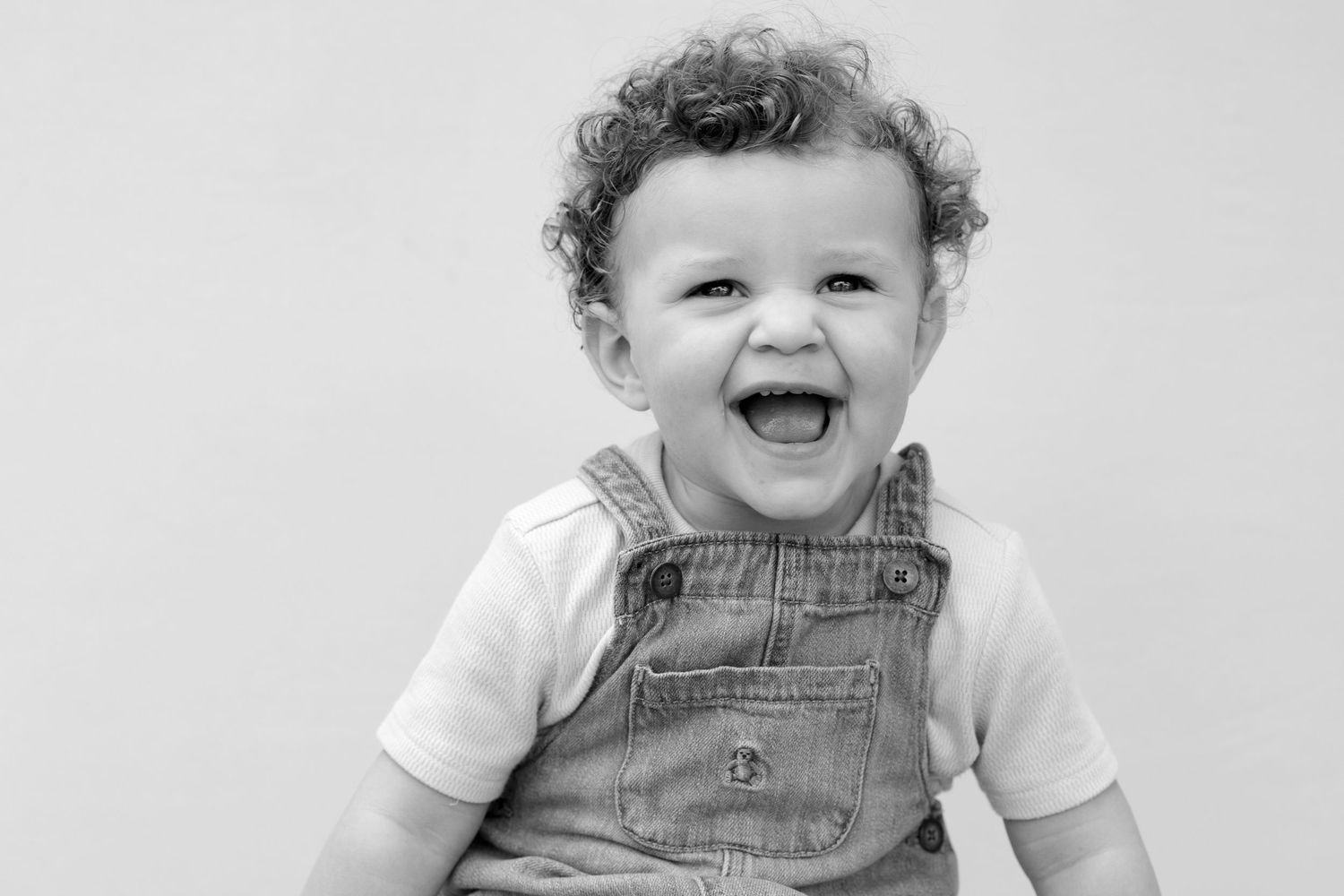 A happy baby wearing denim overalls and a white t-shirt smiles brightly while sitting against a light background.