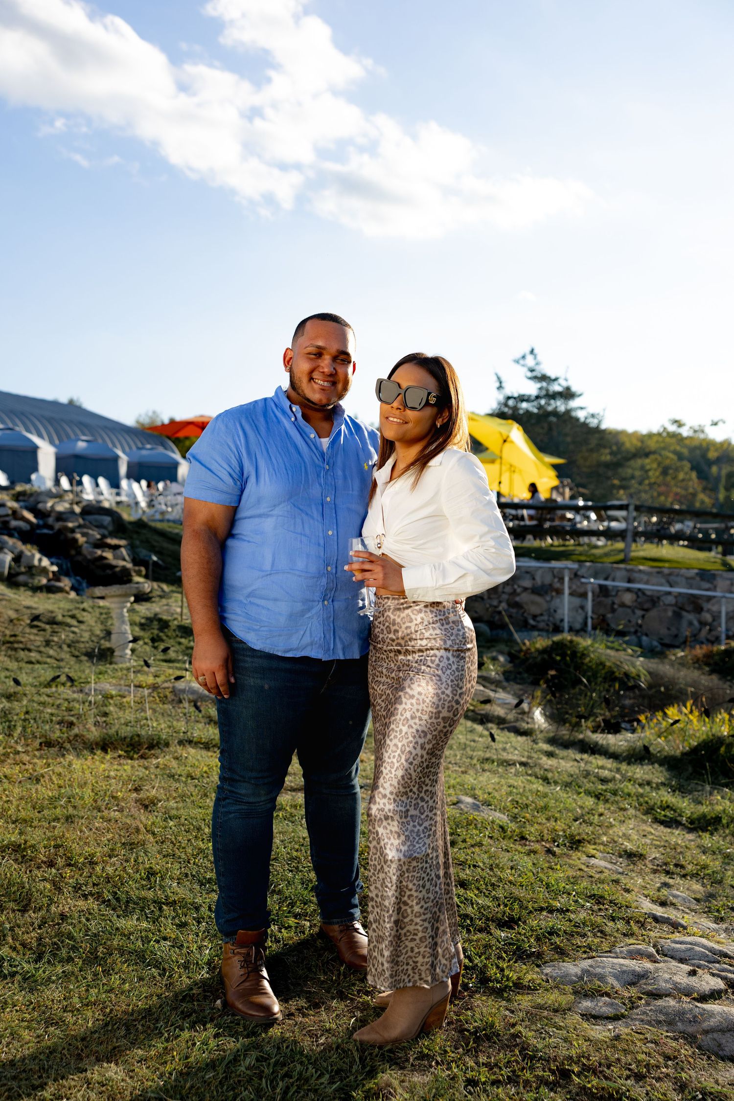 A couple shares a romantic moment on a grassy hillside during golden hour, wearing blue and sparkly outfits.