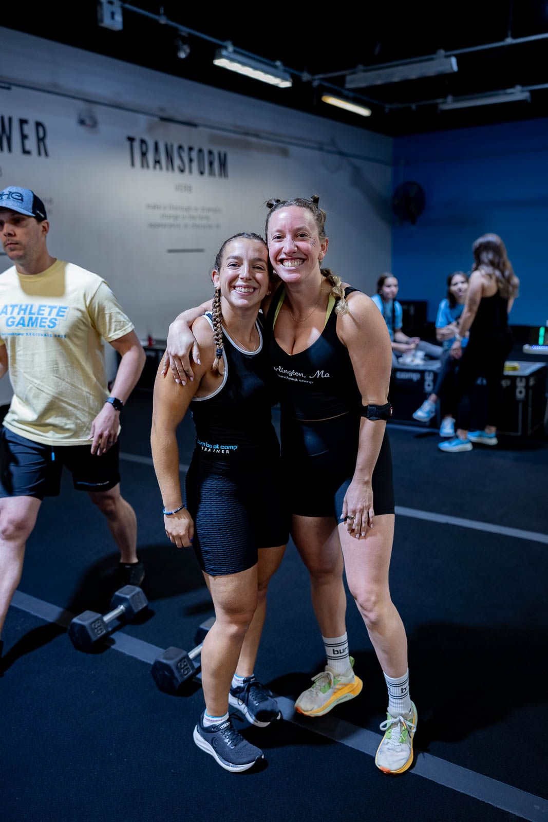 Two athletes in black workout attire smile together at a fitness center with training equipment visible in background.