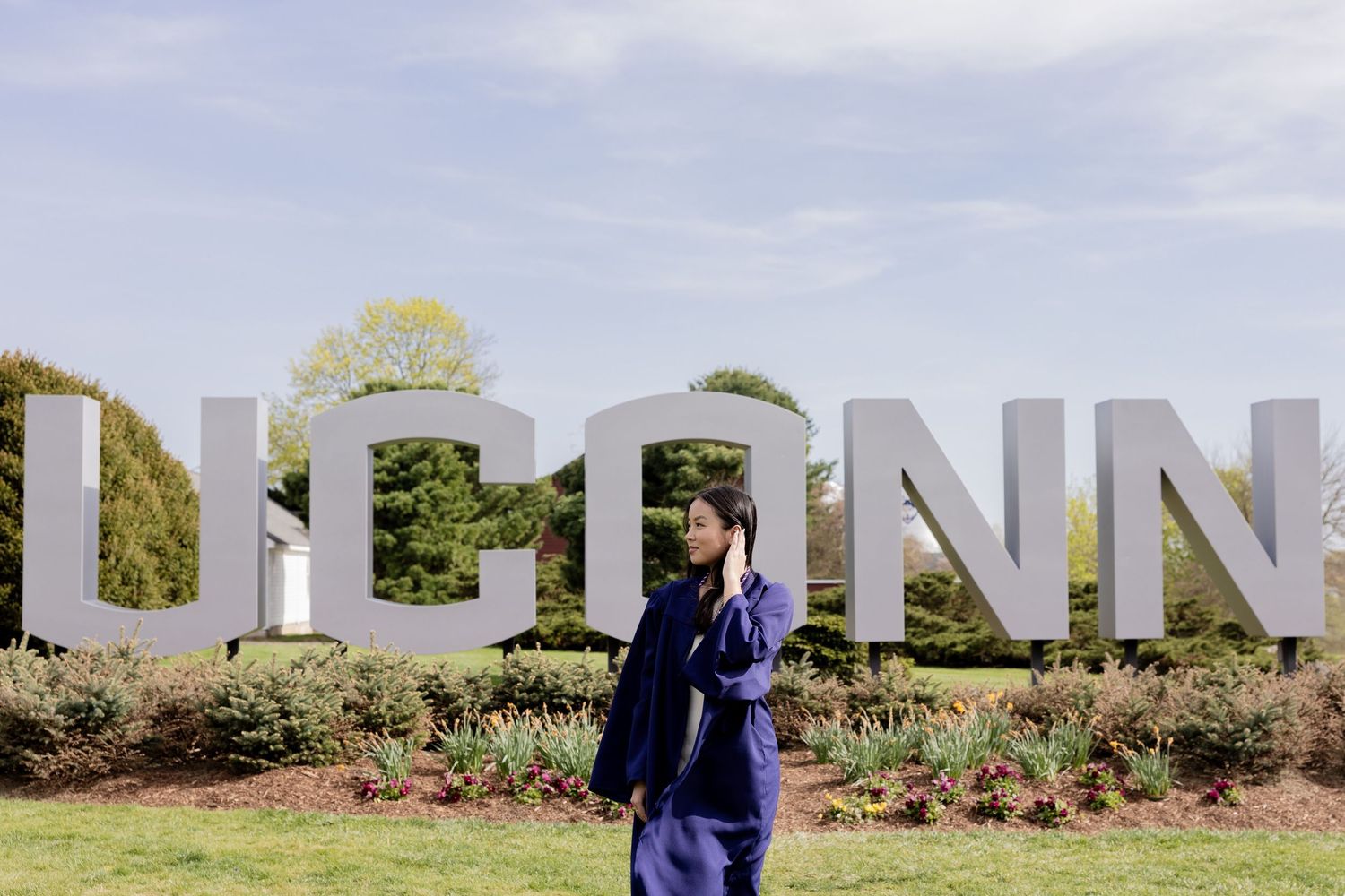 Large white UConn letters create a landmark sign on campus grounds with landscaped gardens and trees in the background.