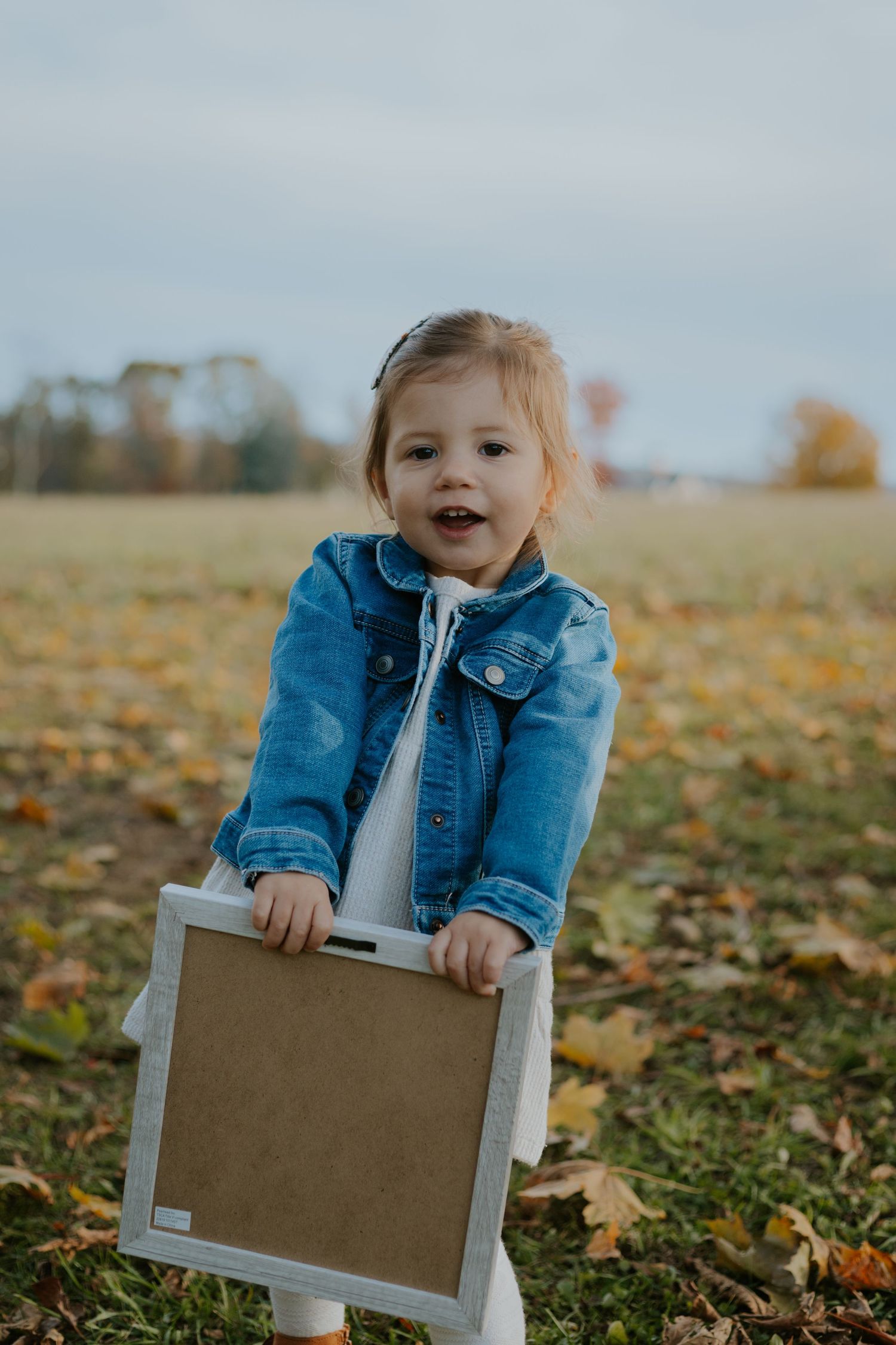 A toddler in a denim jacket holds a brown framed board while standing in fallen leaves.