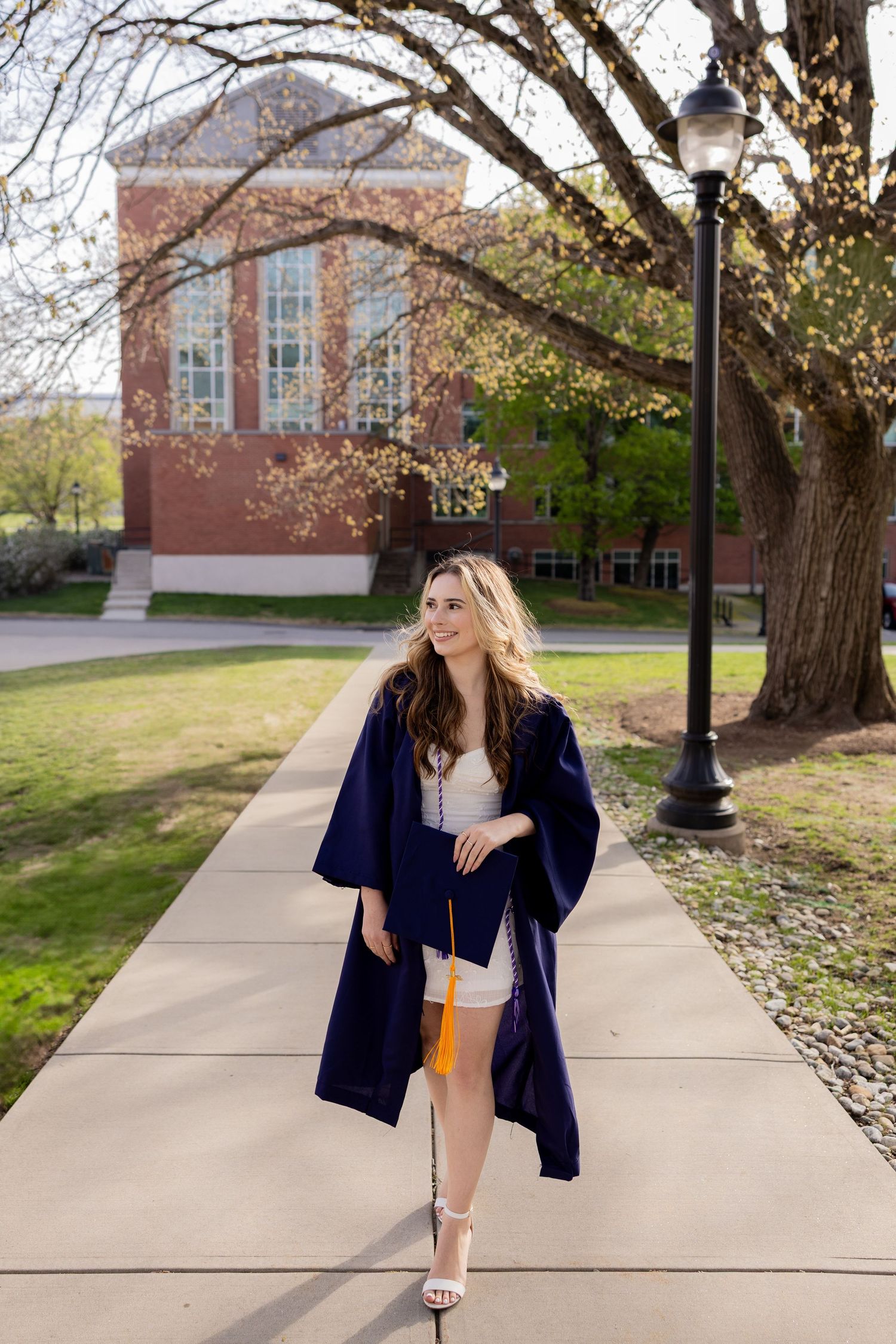 Graduate walks along tree-lined campus path wearing navy blue cap and gown with white dress under bright spring day.