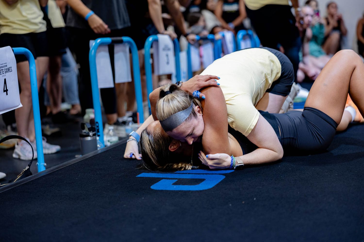 Two competitors grapple during an intense wrestling match on a black mat with blue markings and spectators watching.