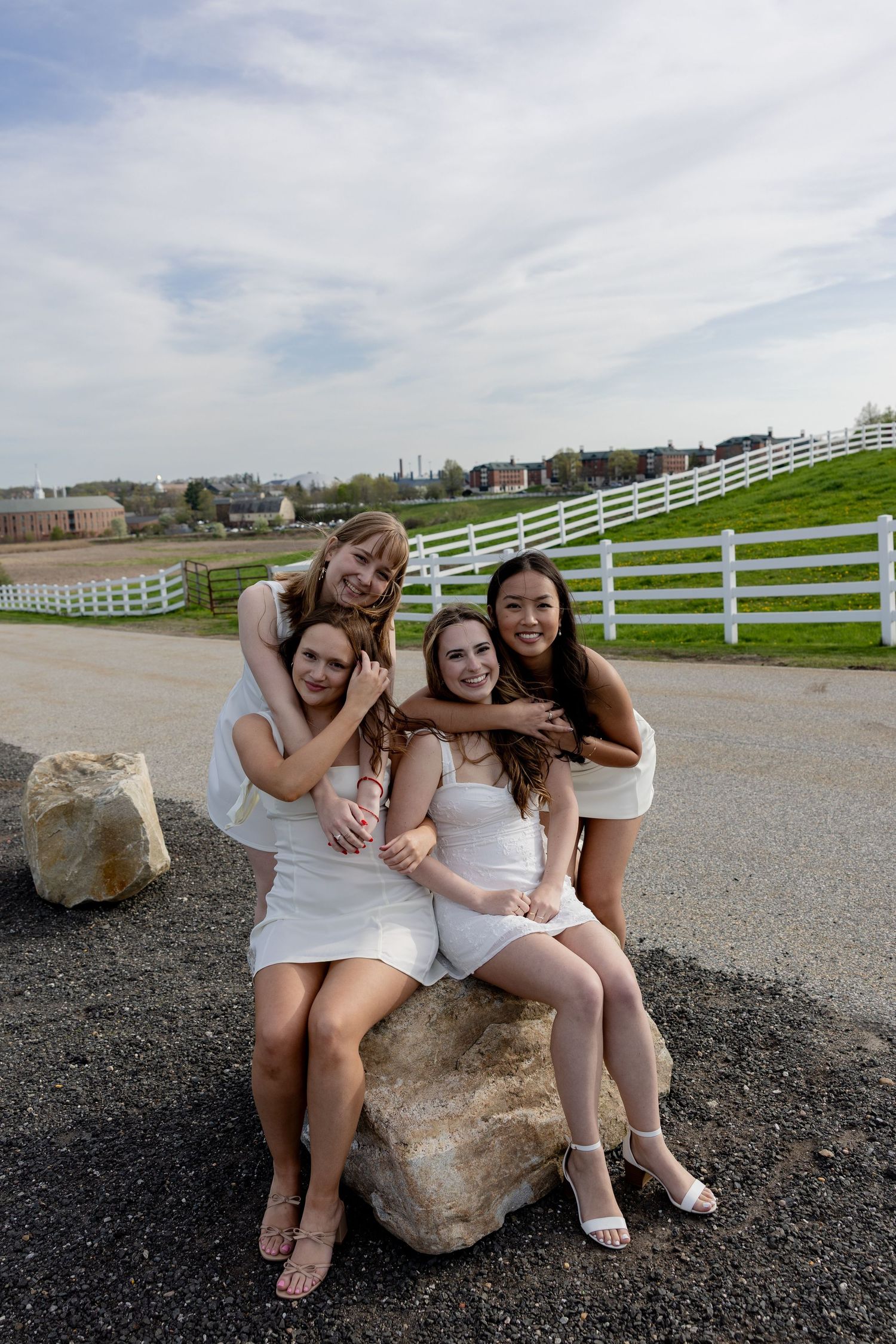 A group sits together on a large rock outdoors with white fencing and green fields in the background.