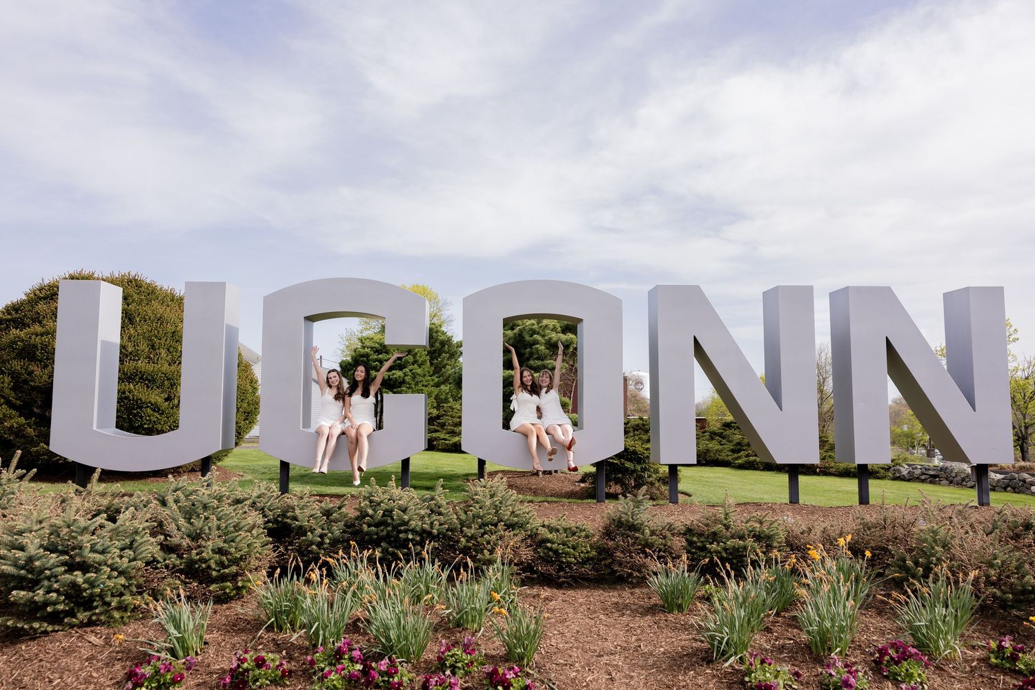 People stand by large white UCONN letters outdoors against a cloudy sky and landscaping.
