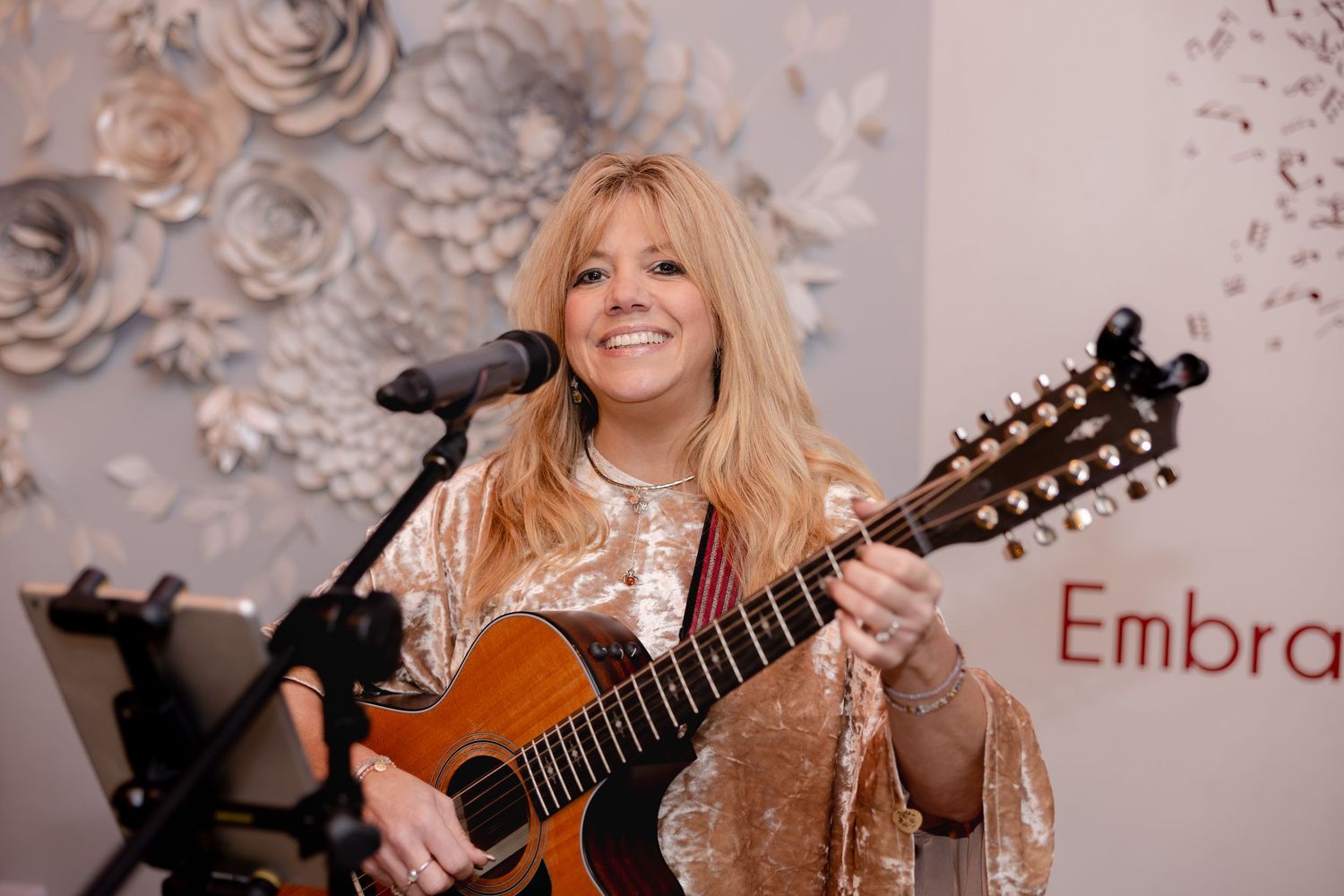 Musician with blonde hair plays acoustic guitar while smiling in front of textured wall background.