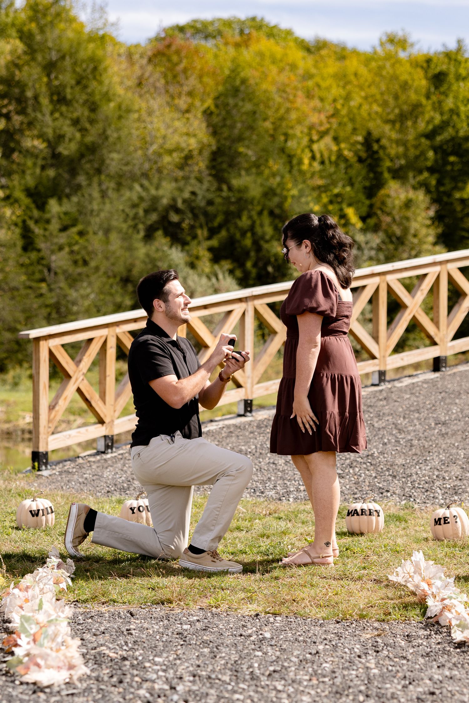 A couple sharing a special moment on a gravel path with wooden railings and autumn foliage in the background.