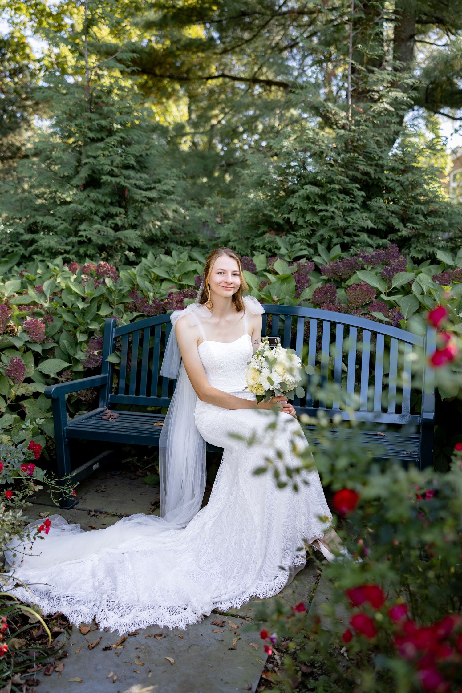 A bride in a white lace gown poses gracefully on a blue garden bench surrounded by blooming roses and greenery.