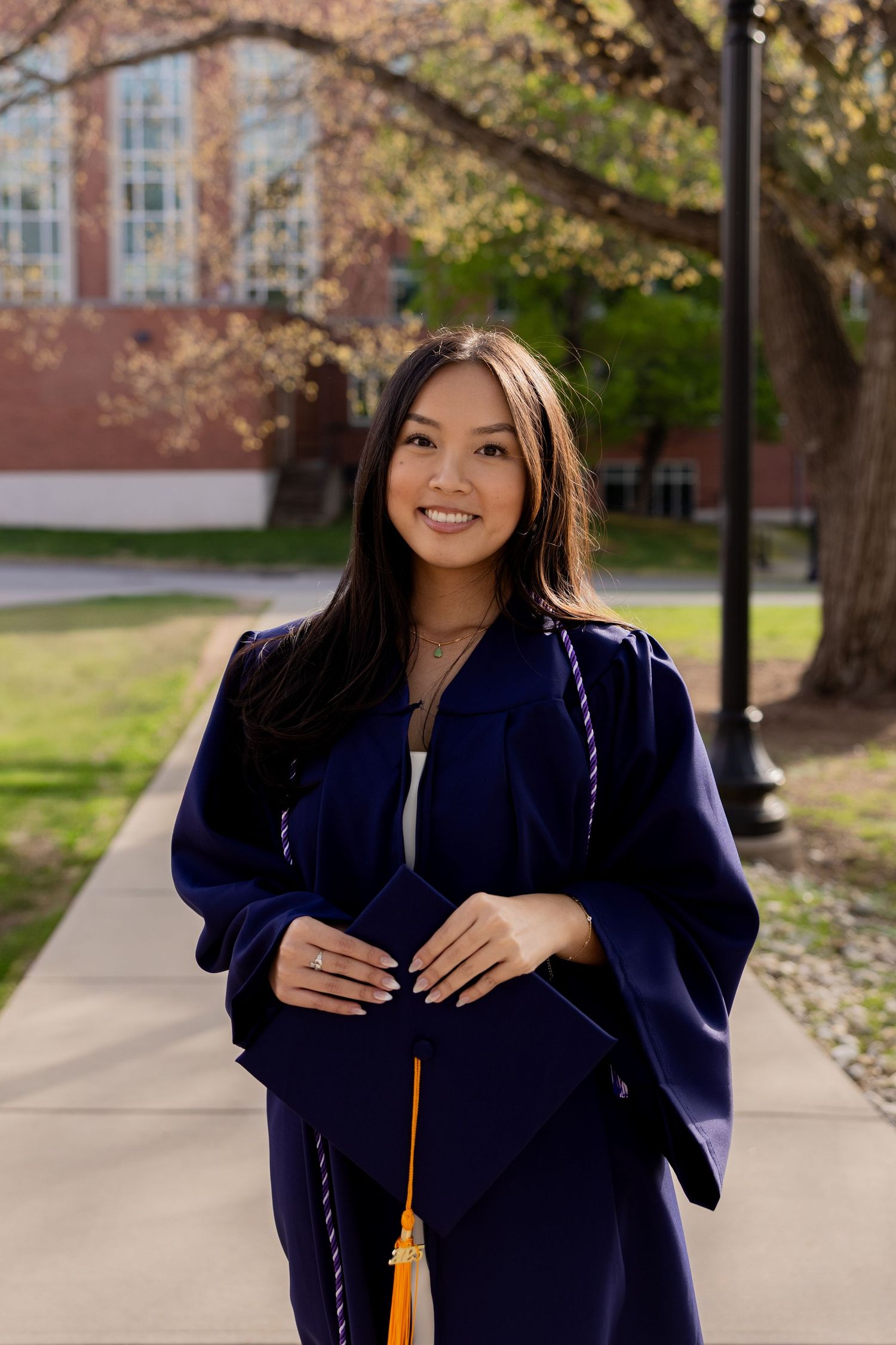Graduate wearing traditional navy blue graduation gown and honor cord stands smiling on university campus pathway.