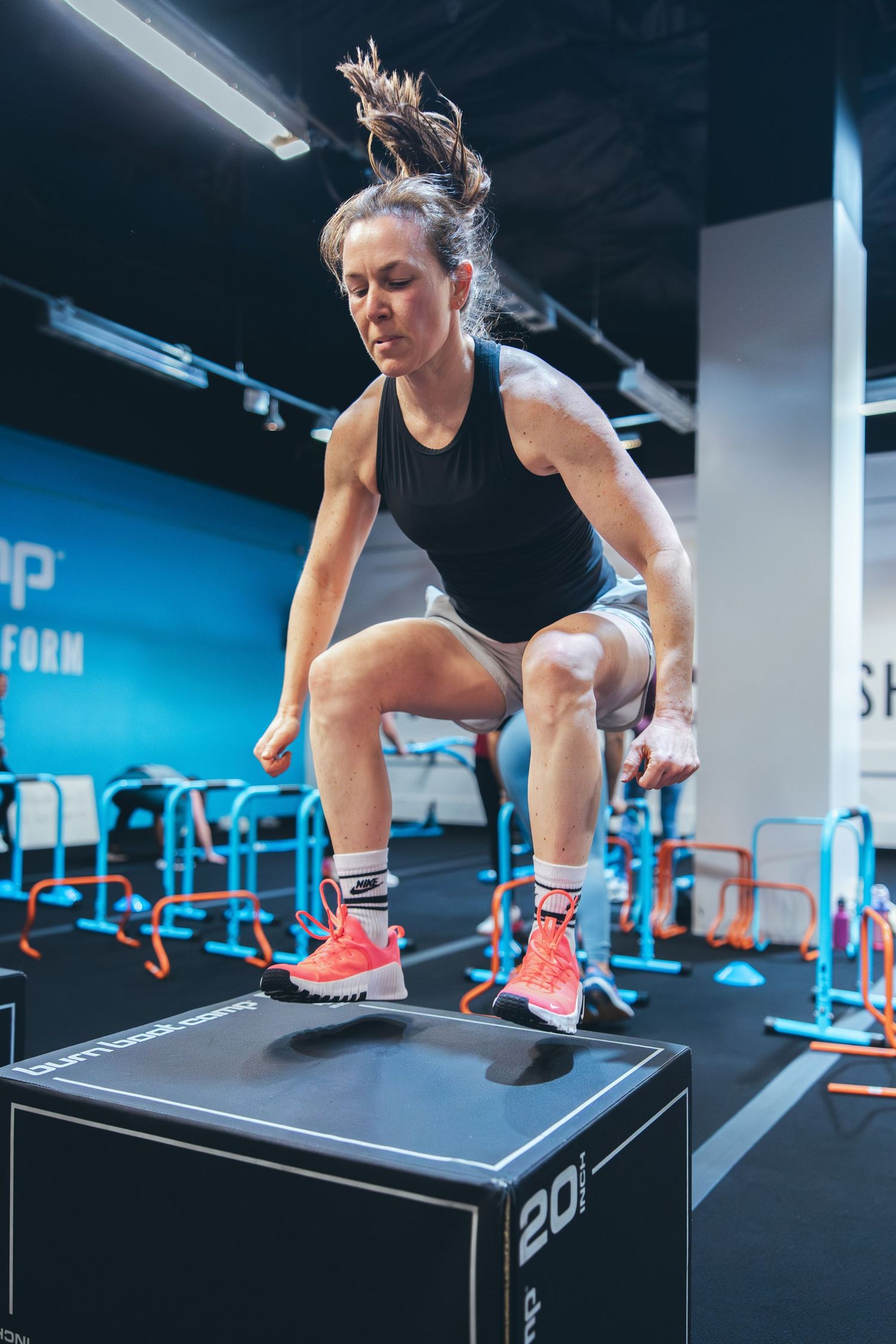 An athlete performs a box jump exercise on a black plyo box in a modern gym setting.