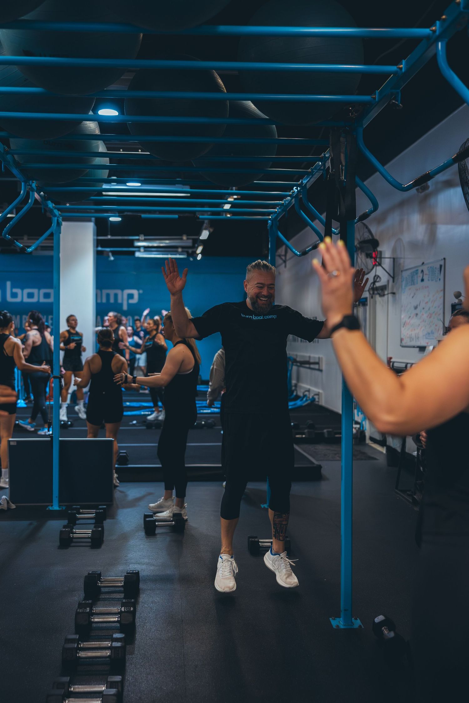Fitness class participants celebrating during a group workout session in a blue-lit gym with dumbbells on the floor.