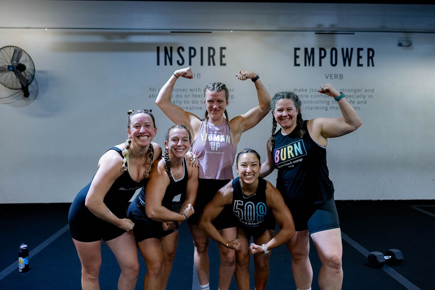 A group of athletic women posing together and flexing their muscles in front of an 'Inspire Empower' wall at a gym.