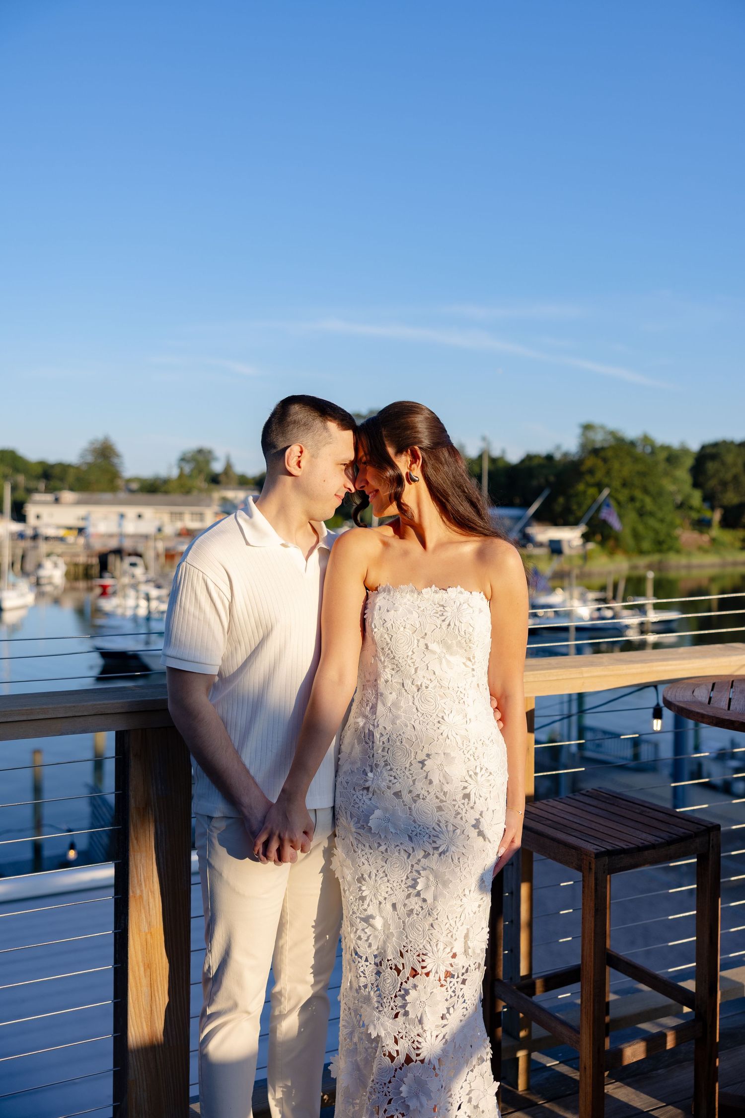 A couple shares a romantic moment at sunset on a waterfront deck with boats and docks visible in the background.