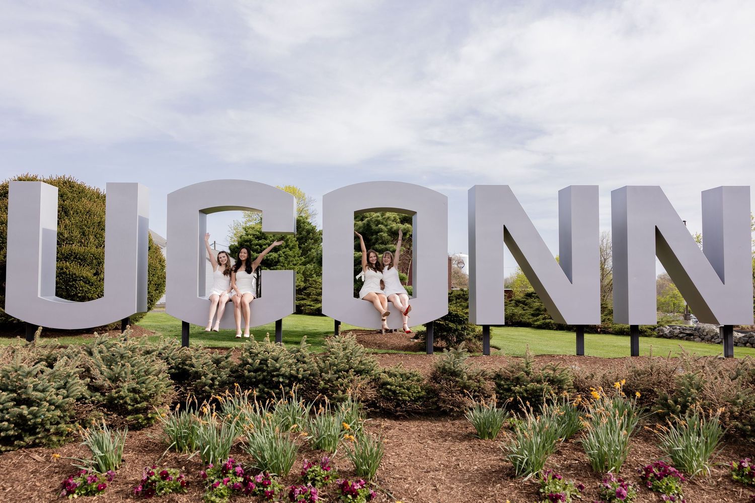 Three people pose with large white UCONN letters display outdoors on a landscaped hill against cloudy sky.