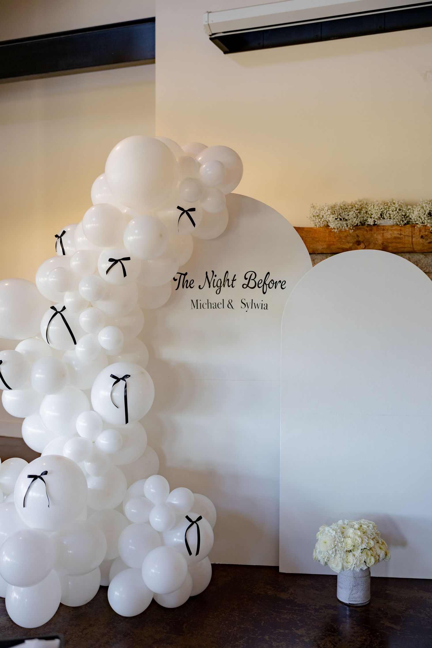 An elegant white balloon arch with black bows creates a wedding backdrop against a cream-colored wall.