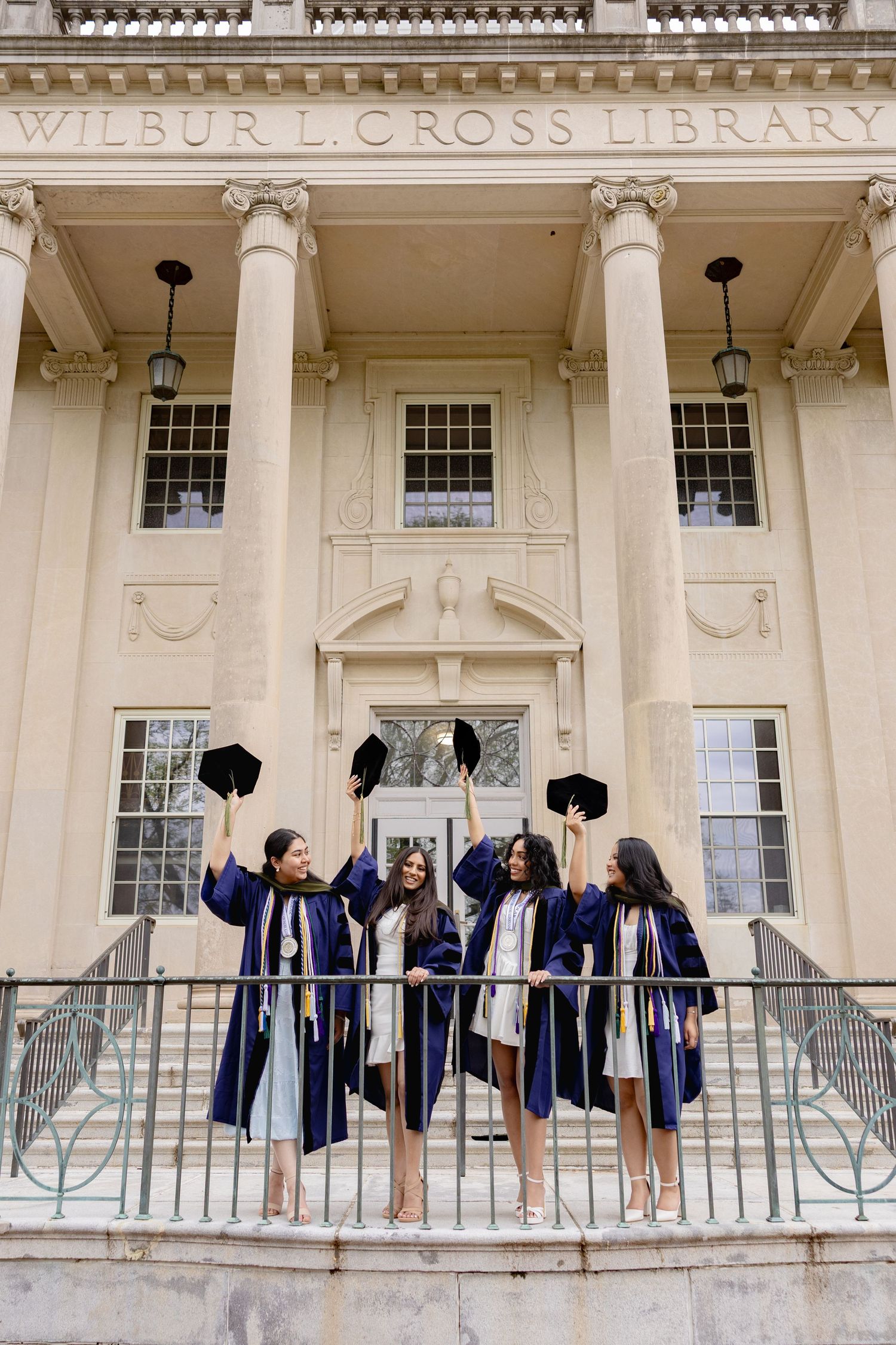 Graduates in blue caps and gowns celebrate on the steps of Wilbur L Cross Library with raised arms.