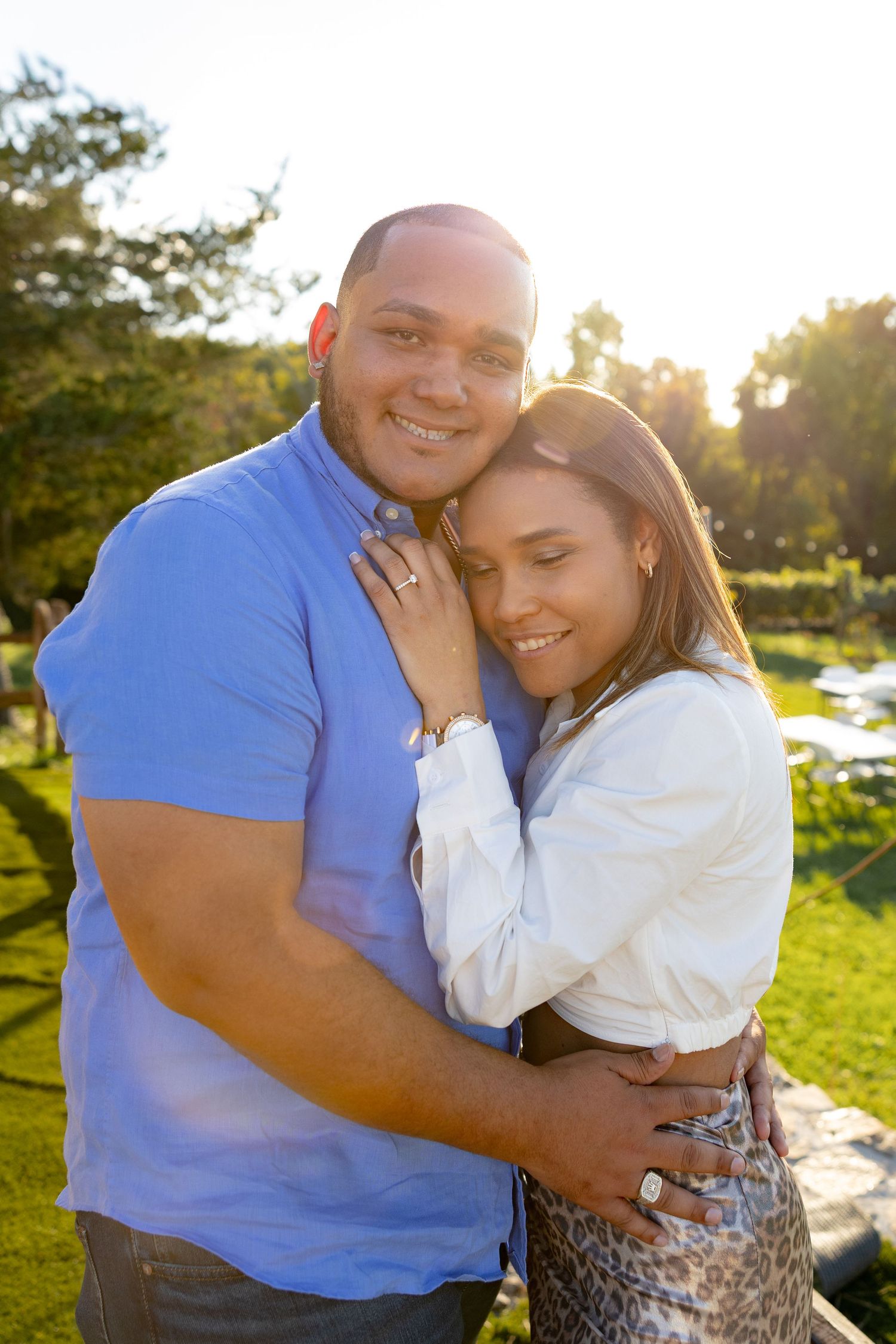 Couple embracing in warm sunlight with green landscape and parking area visible in background.