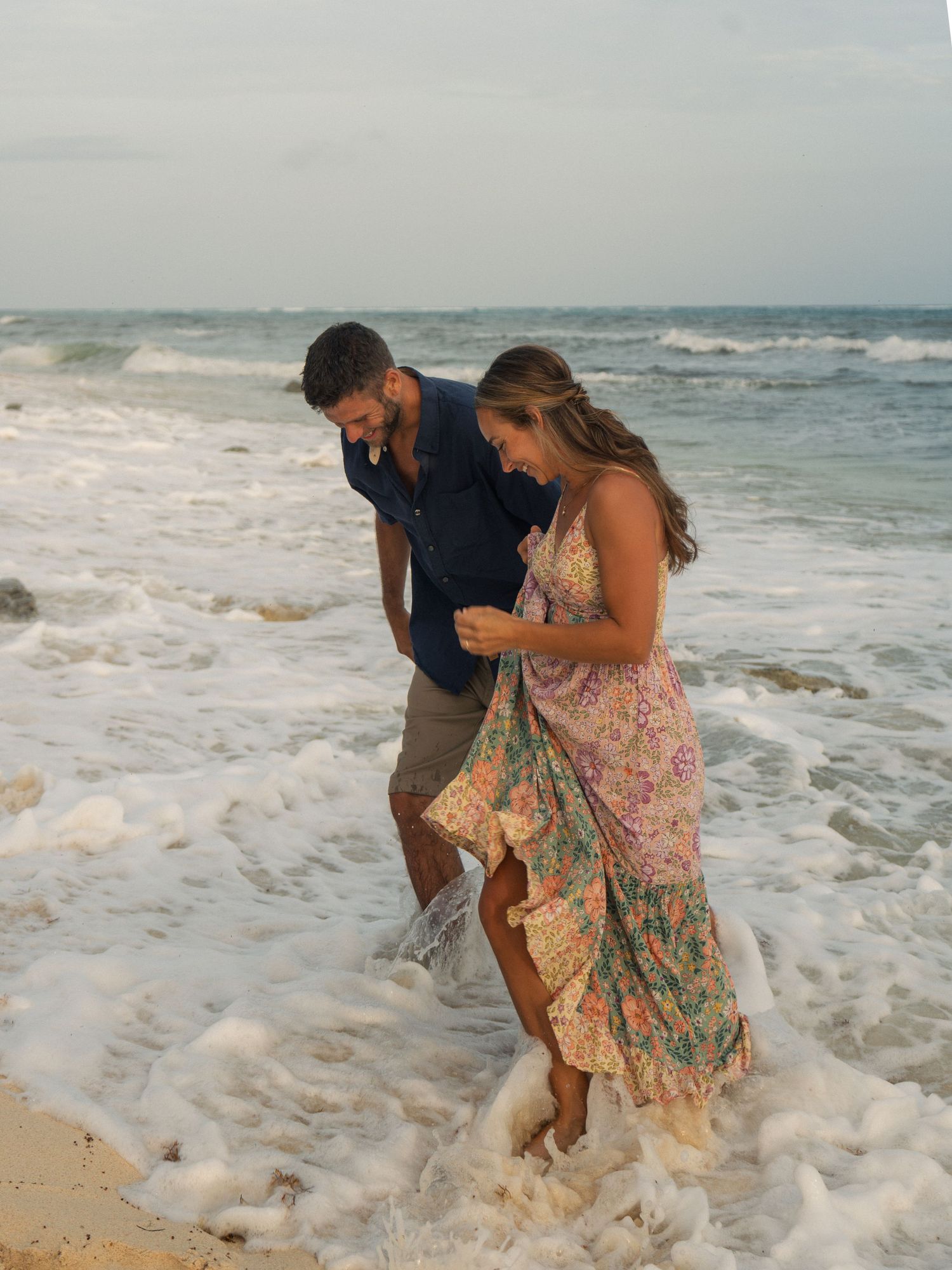 Couple laughing as ocean waves wash over their feet on a sandy beach at sunset.