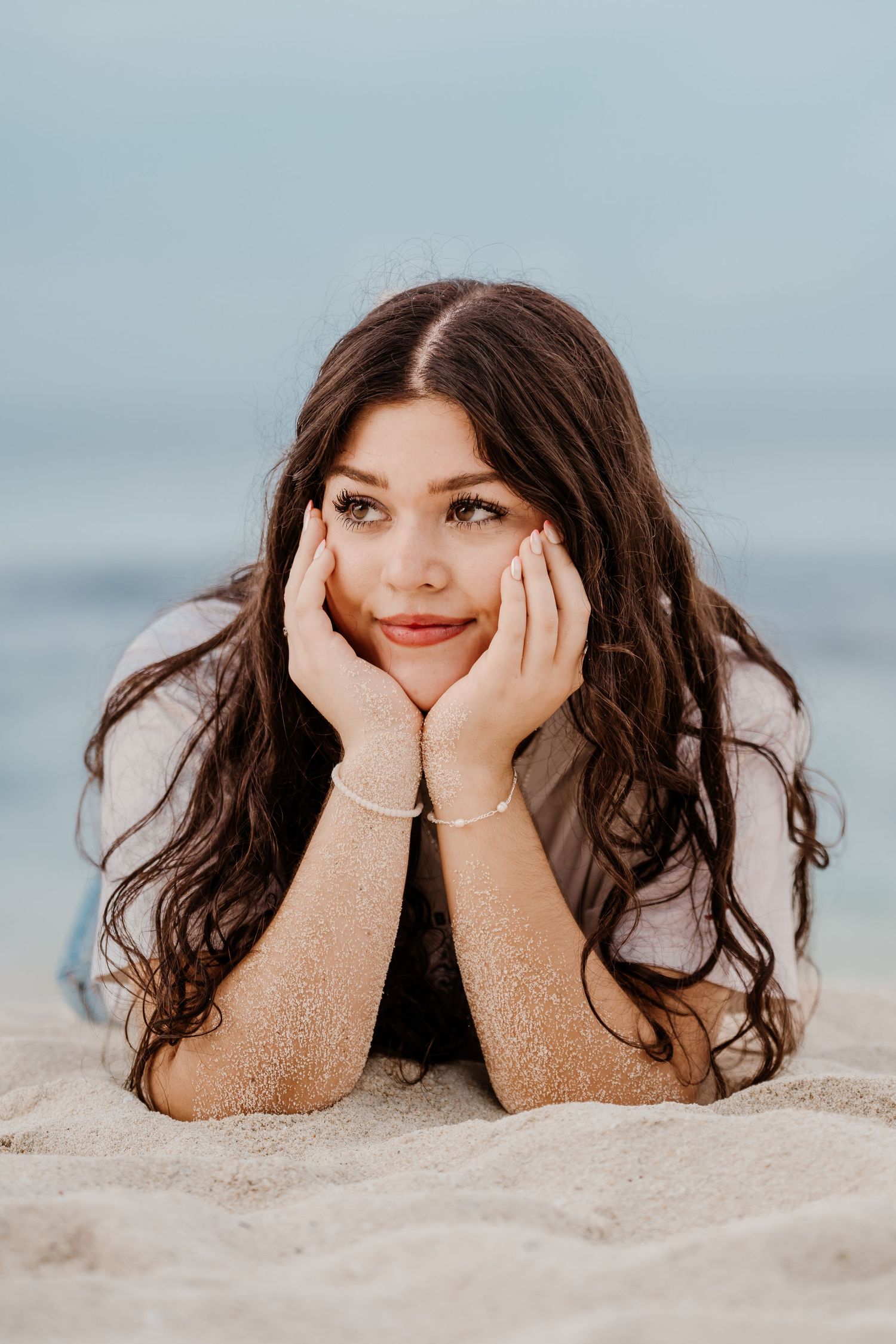 Person lying on sandy beach with hands under chin in relaxed pose.