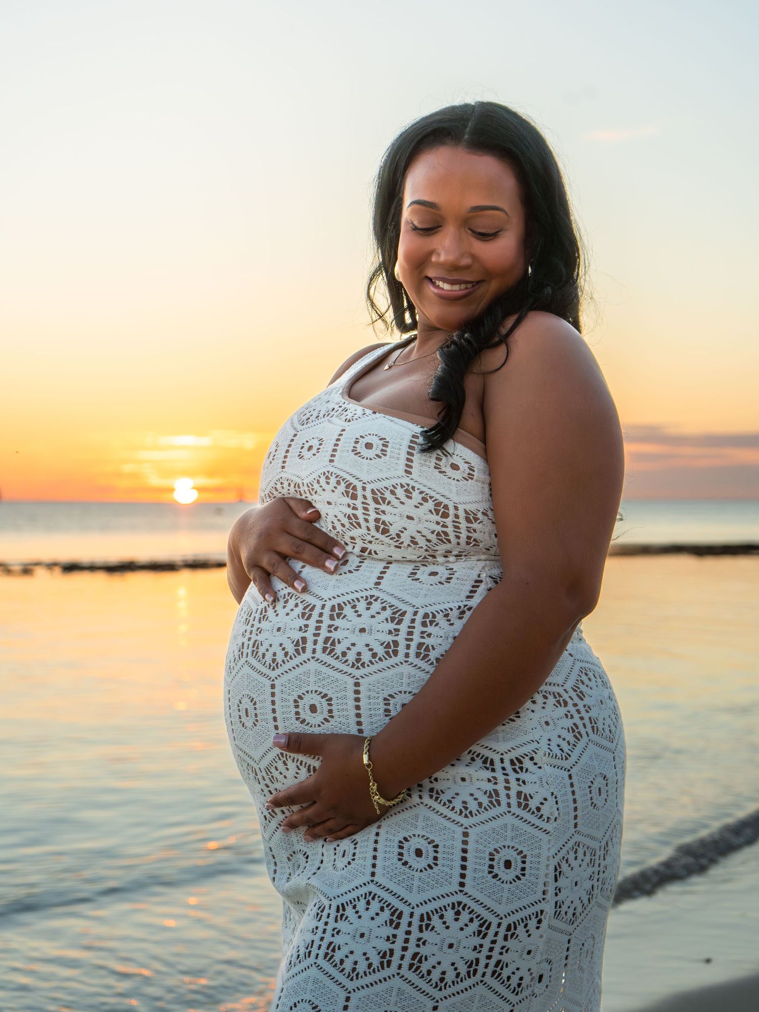Pregnant woman in white lace dress cradling her baby bump at sunset by the ocean.