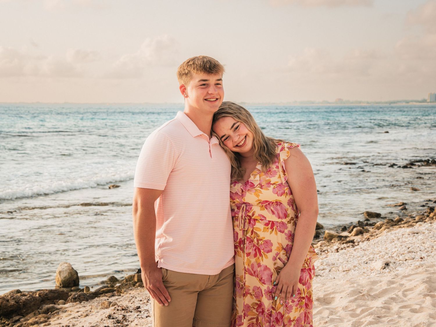 A couple smiles together on a sandy beach at sunset, the woman in a floral dress and man in a pink polo.
