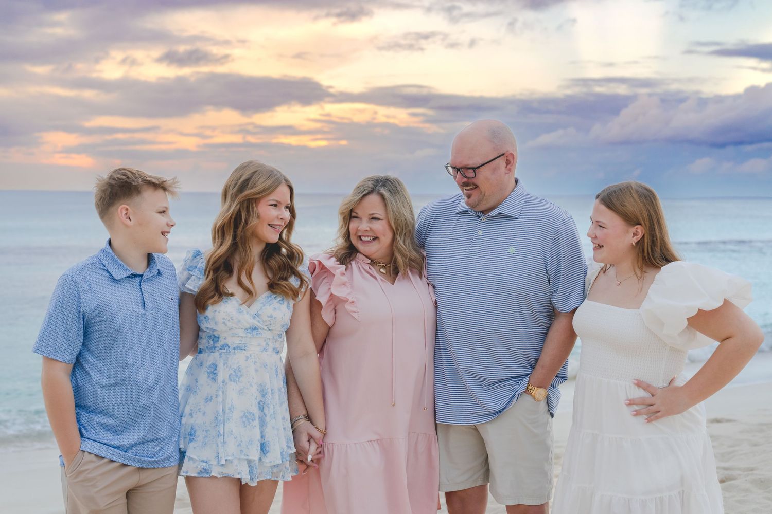 A family wearing coordinated blue and pink outfits poses together on a beach at sunset.