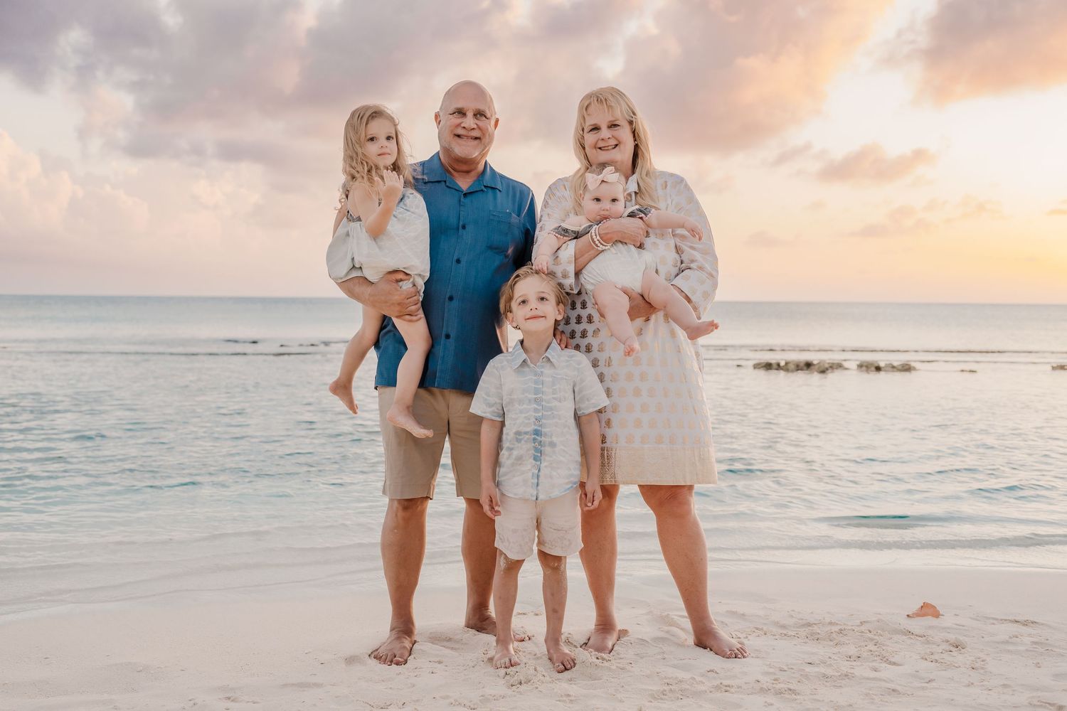 A family enjoys a playful moment together on a sandy beach at sunset with the ocean in the background.