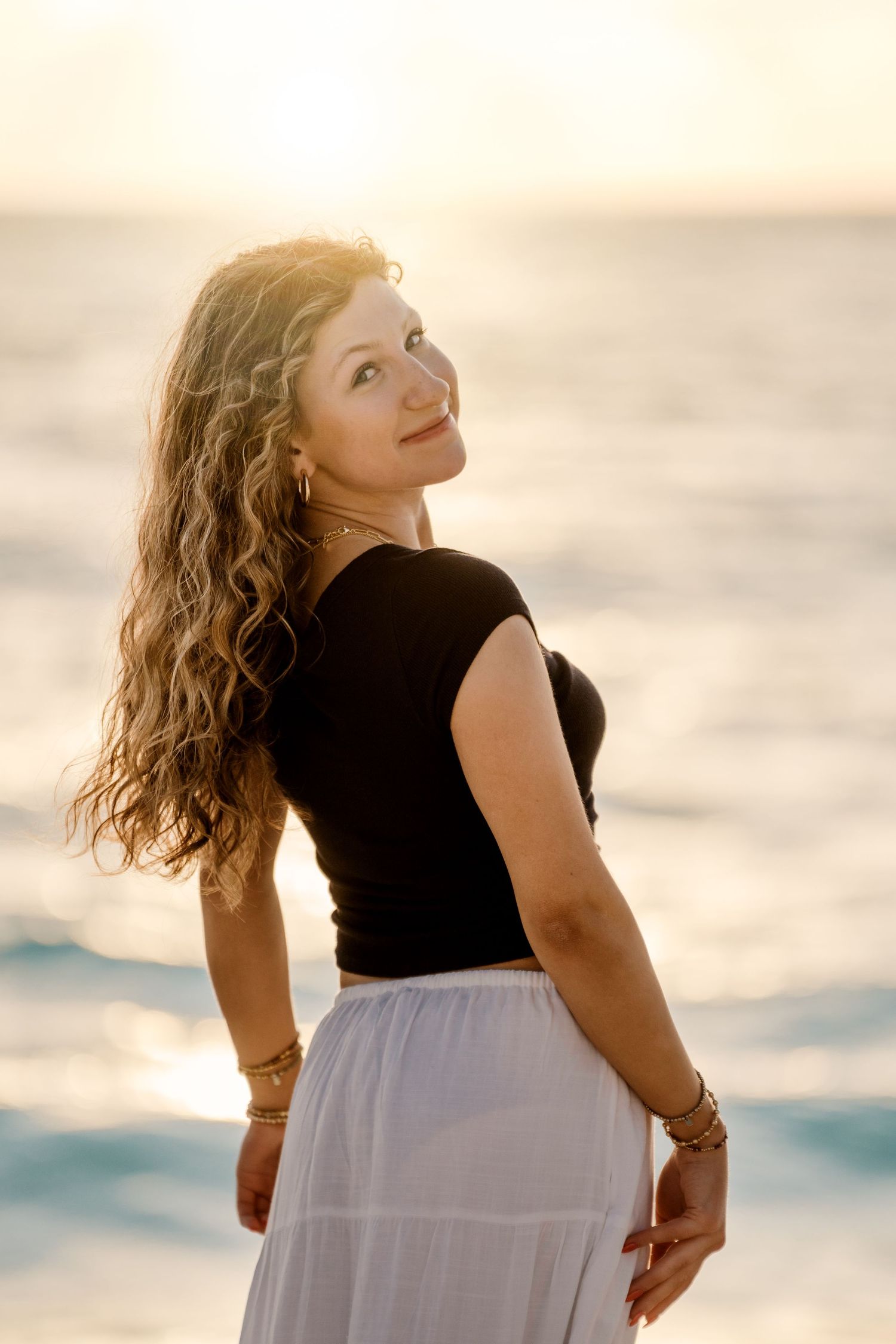 A person in a black and white outfit poses on a beach during sunset with waves visible behind.