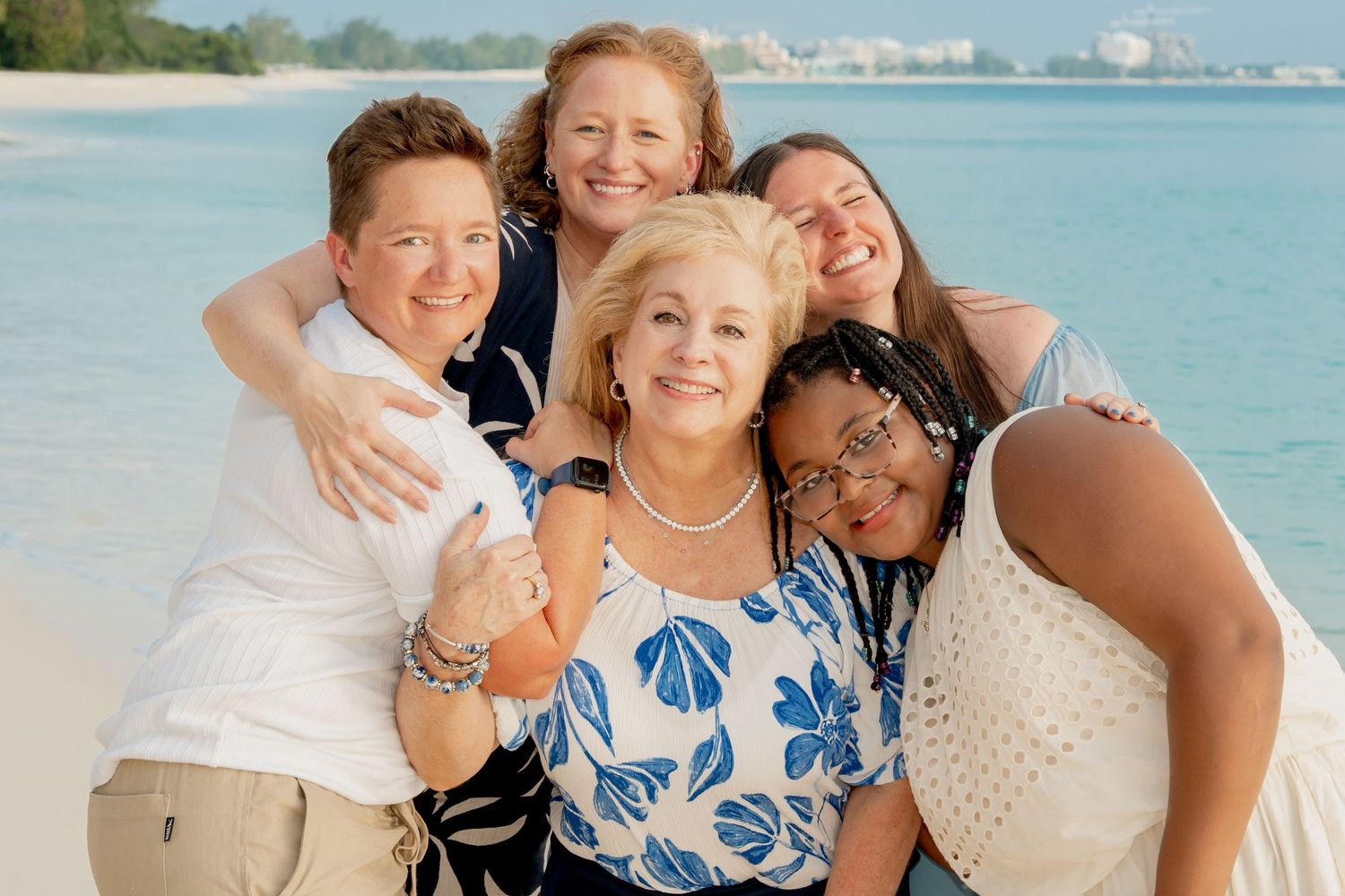 Five women smiling and embracing on a tropical beach with turquoise water in the background.