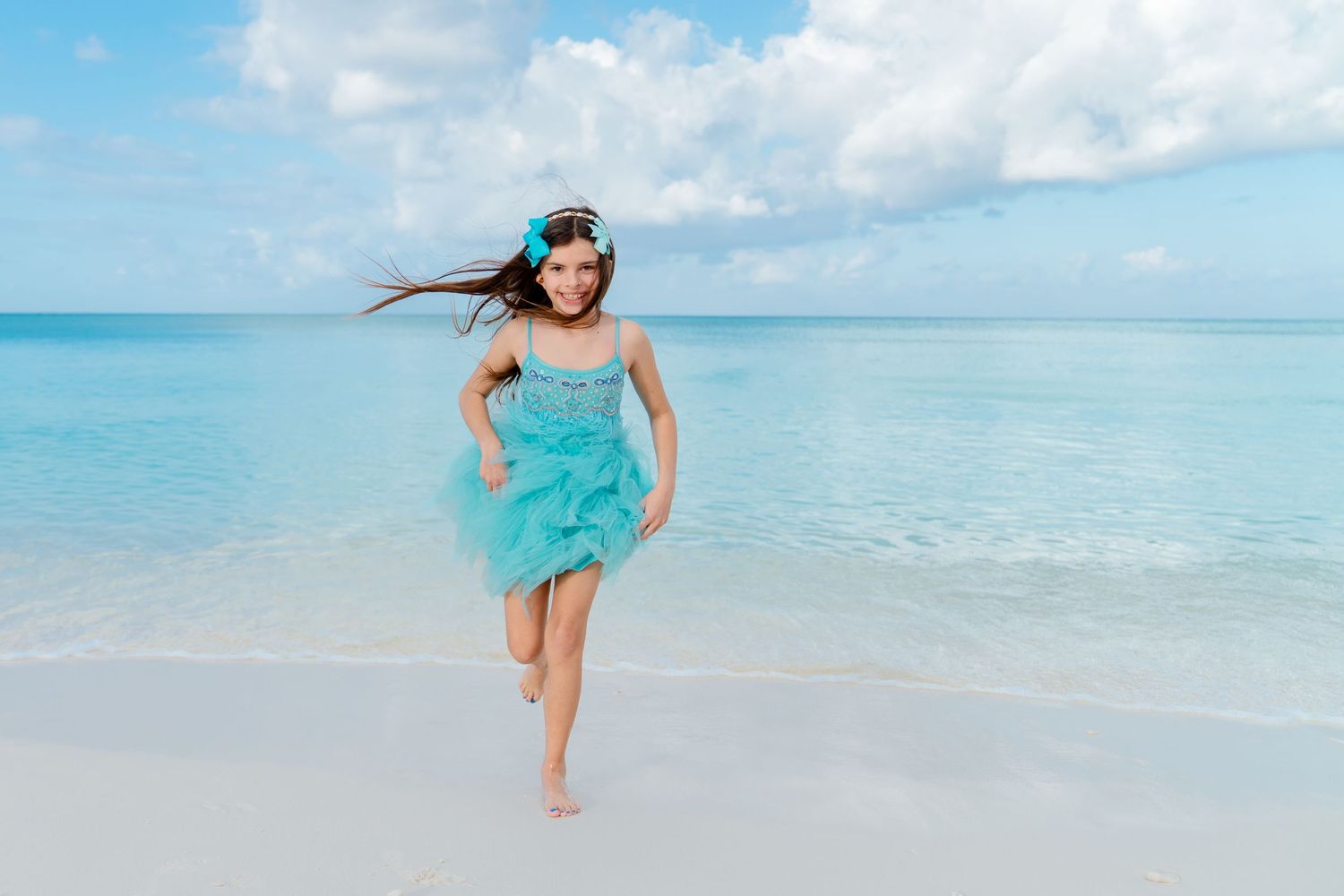 A person in a flowing turquoise dress runs joyfully along a pristine white sand beach with crystal clear waters.