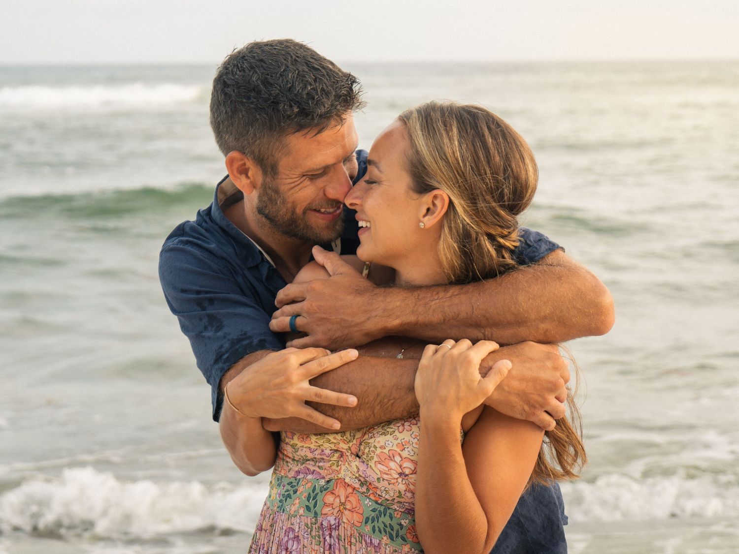 Couple embracing and smiling at each other on a beach with ocean waves in the background.