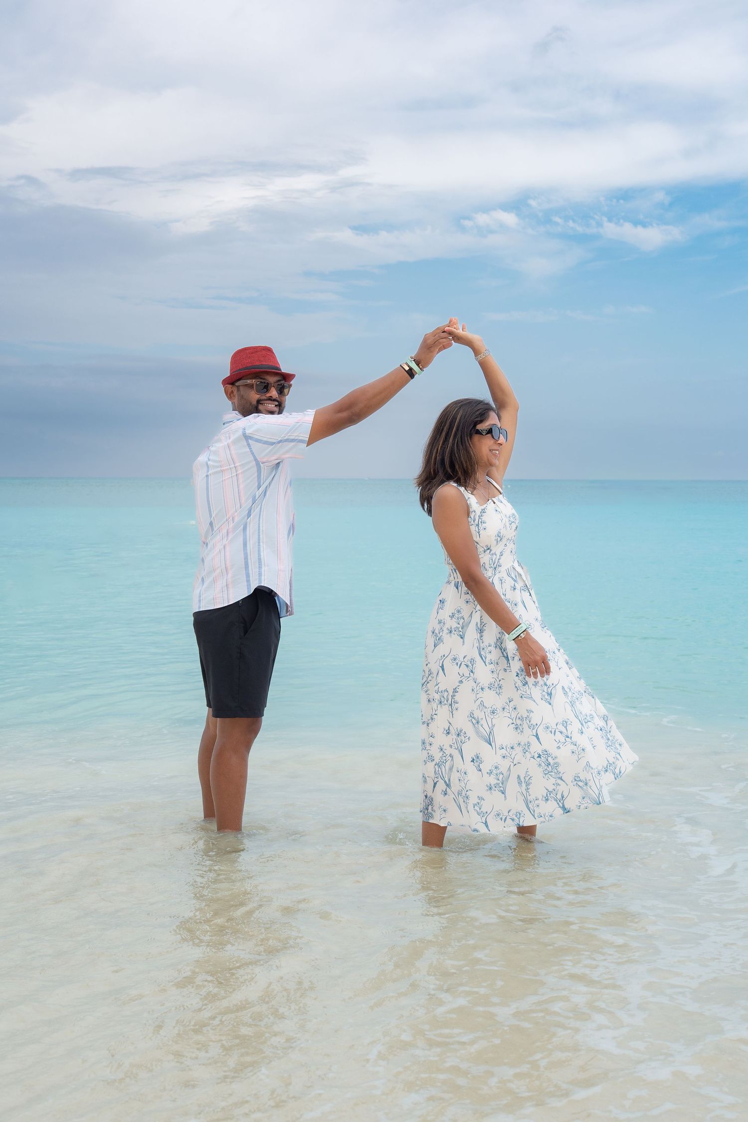 Couple dancing in turquoise ocean water, man in red hat and white shirt, woman in floral dress, tropical beach.