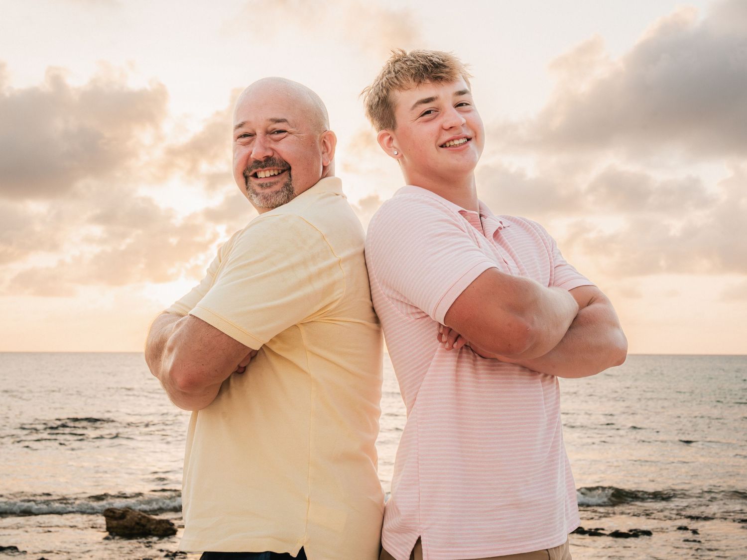 Two men standing back-to-back with arms crossed, smiling on a beach at sunset.