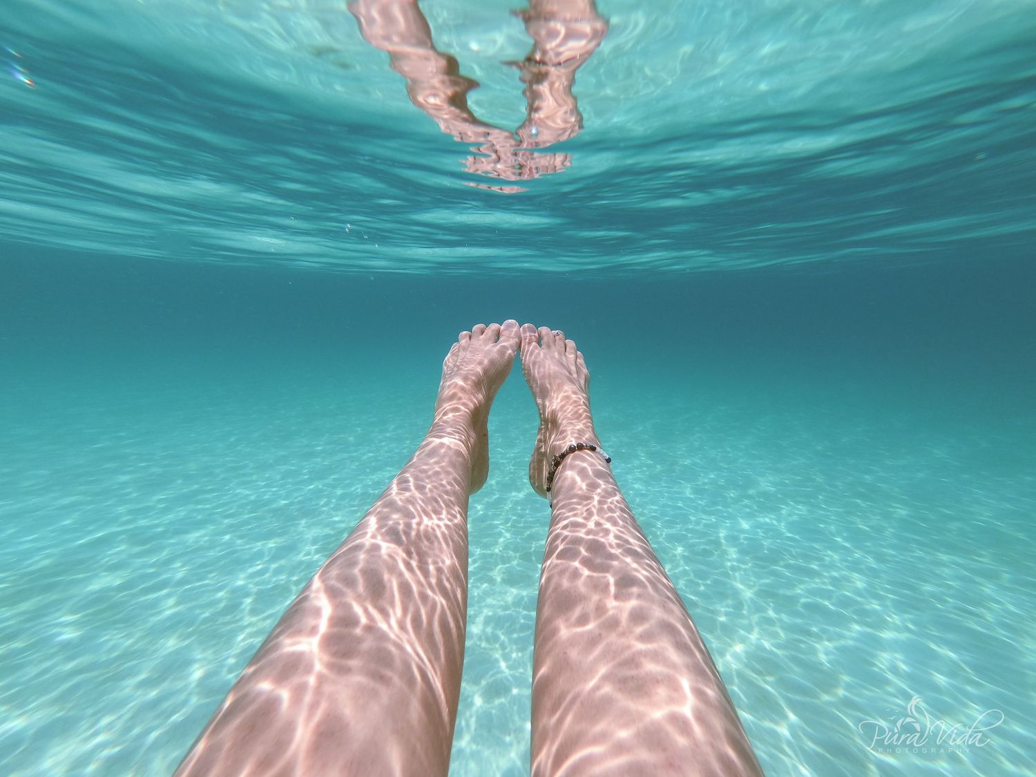 Underwater view of legs dangling in crystal clear turquoise water above sandy ocean floor.