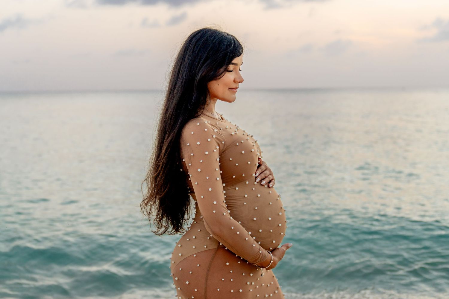 Side view of pregnant woman in beige dress posing against turquoise ocean backdrop.