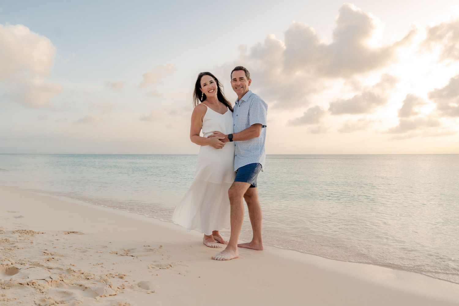 Couple embracing on a tropical beach at sunset, woman in white dress, man in blue shirt and shorts, turquoise ocean backdrop.