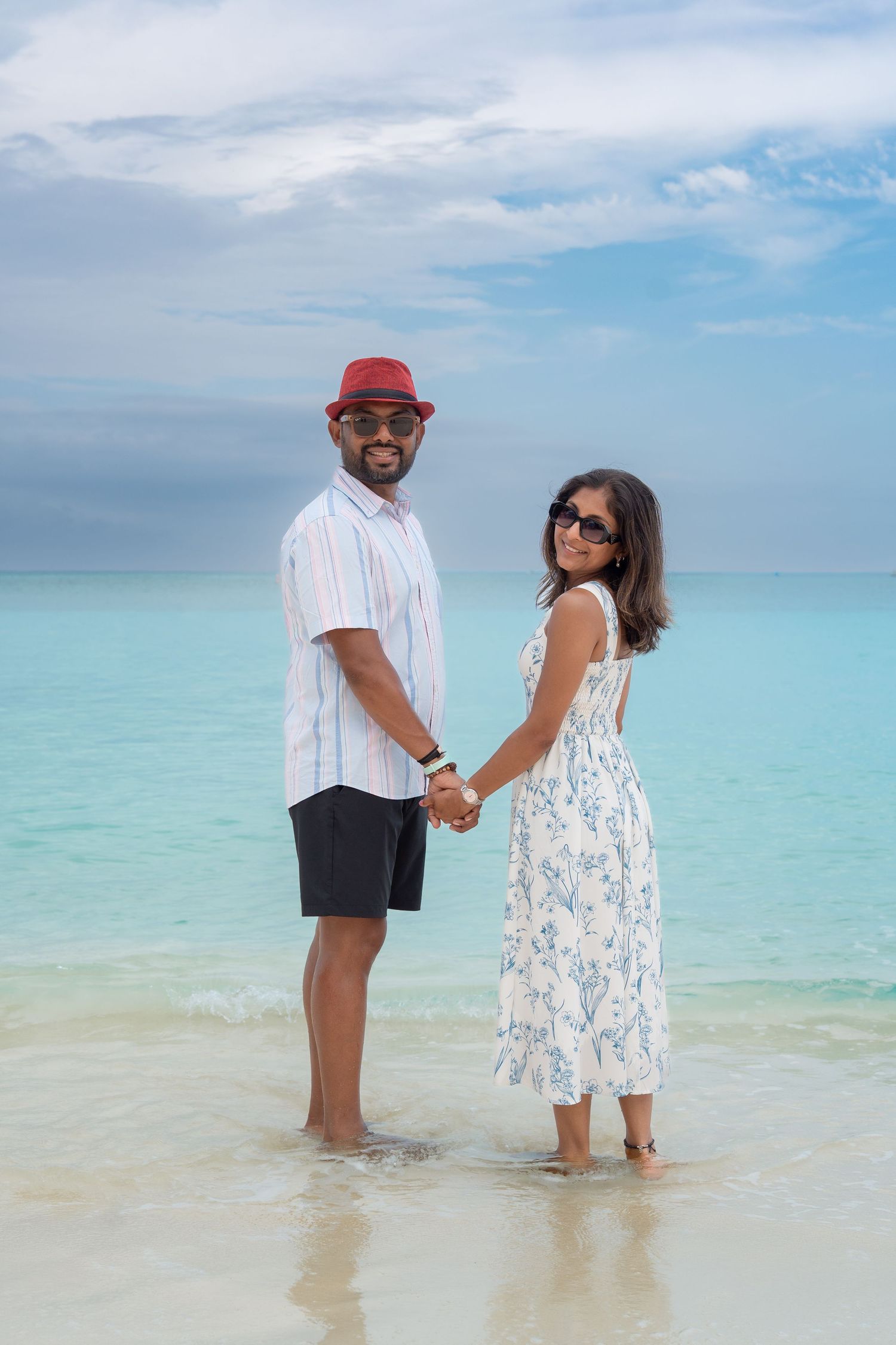 Couple holding hands on a tropical beach, man in red hat and sunglasses, woman in floral dress, turquoise ocean backdrop.