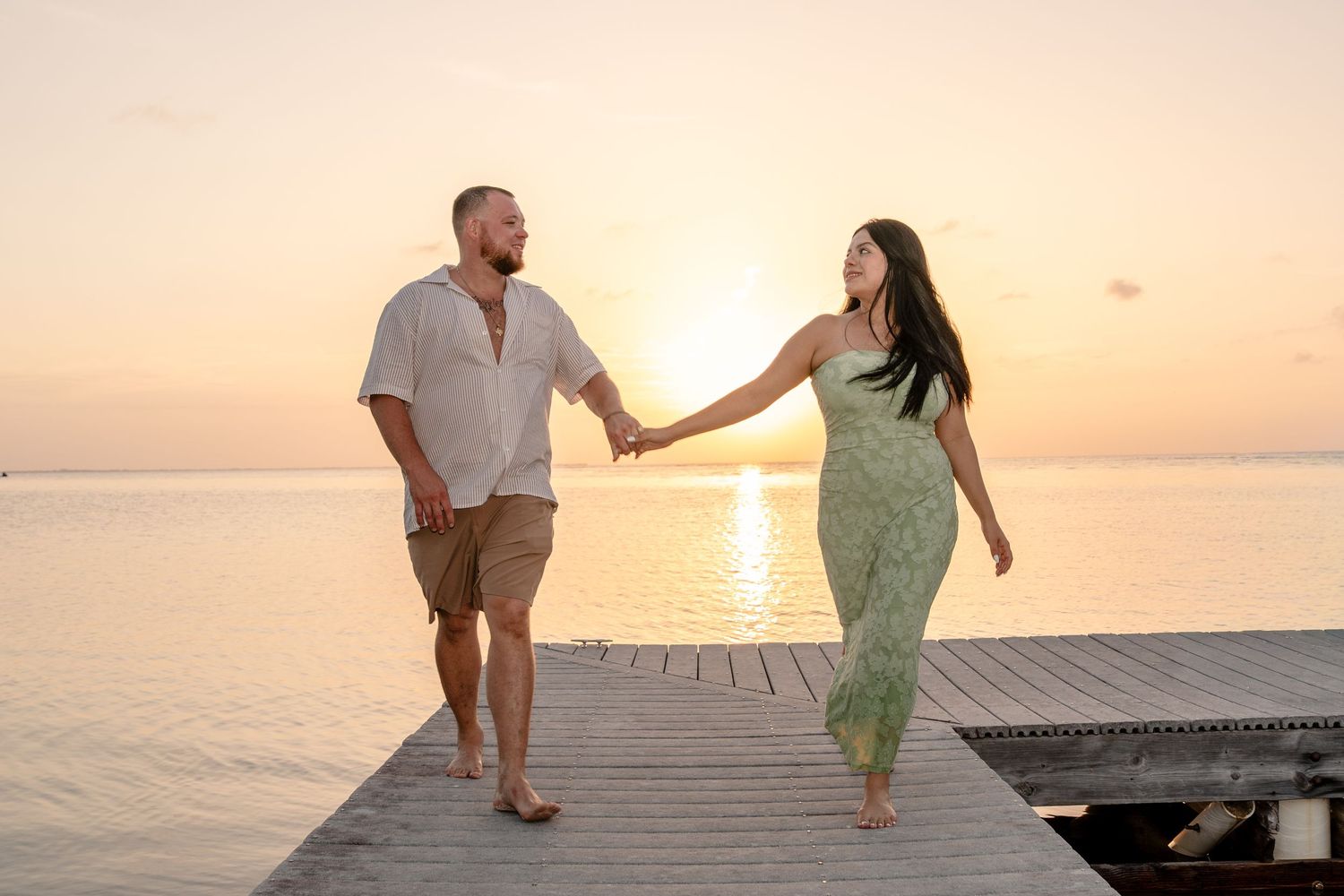 Couple holding hands walking on a wooden pier at sunset over calm ocean waters.