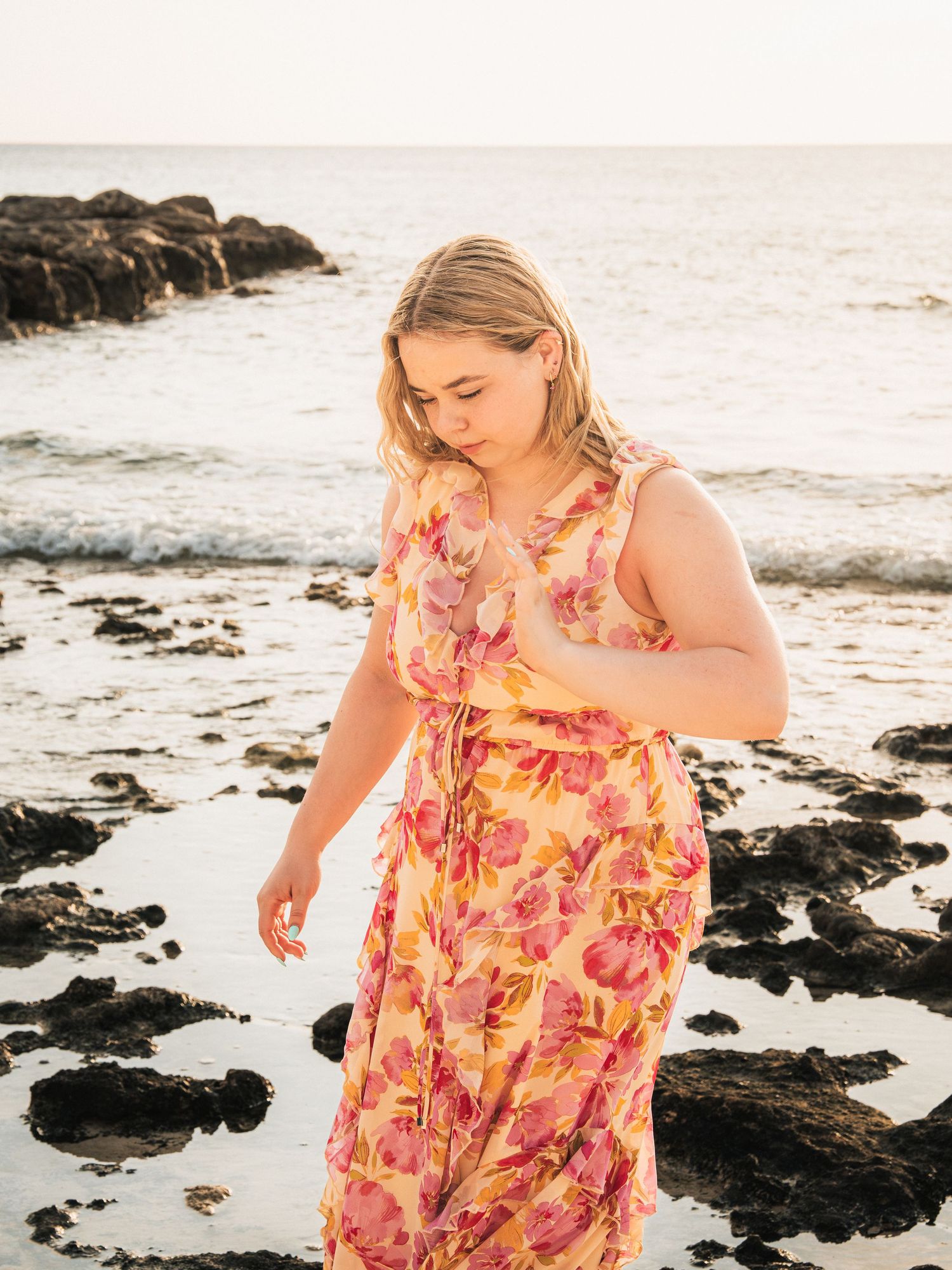 A blonde woman in a vibrant floral maxi dress stands thoughtfully on a rocky beach at golden hour.