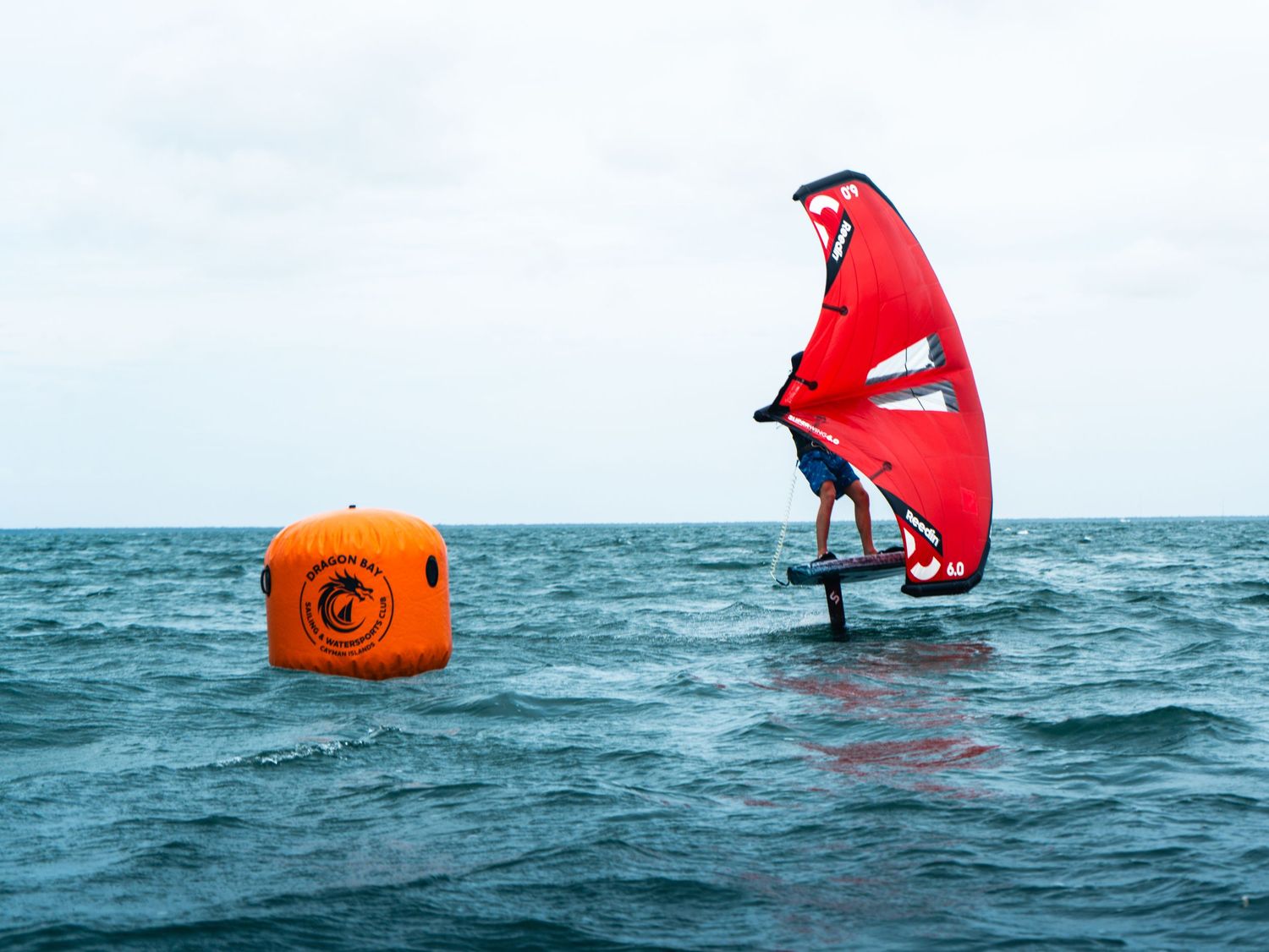 Windsurfer on a hydrofoil glides past an orange buoy marker on open ocean waters.