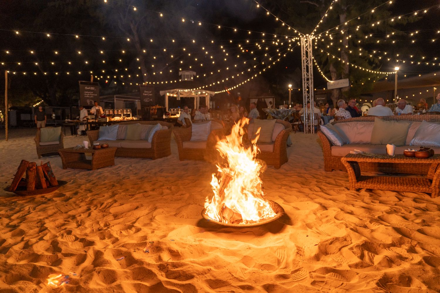 Bonfires illuminate a sandy beach at night with string lights overhead creating a magical atmosphere.