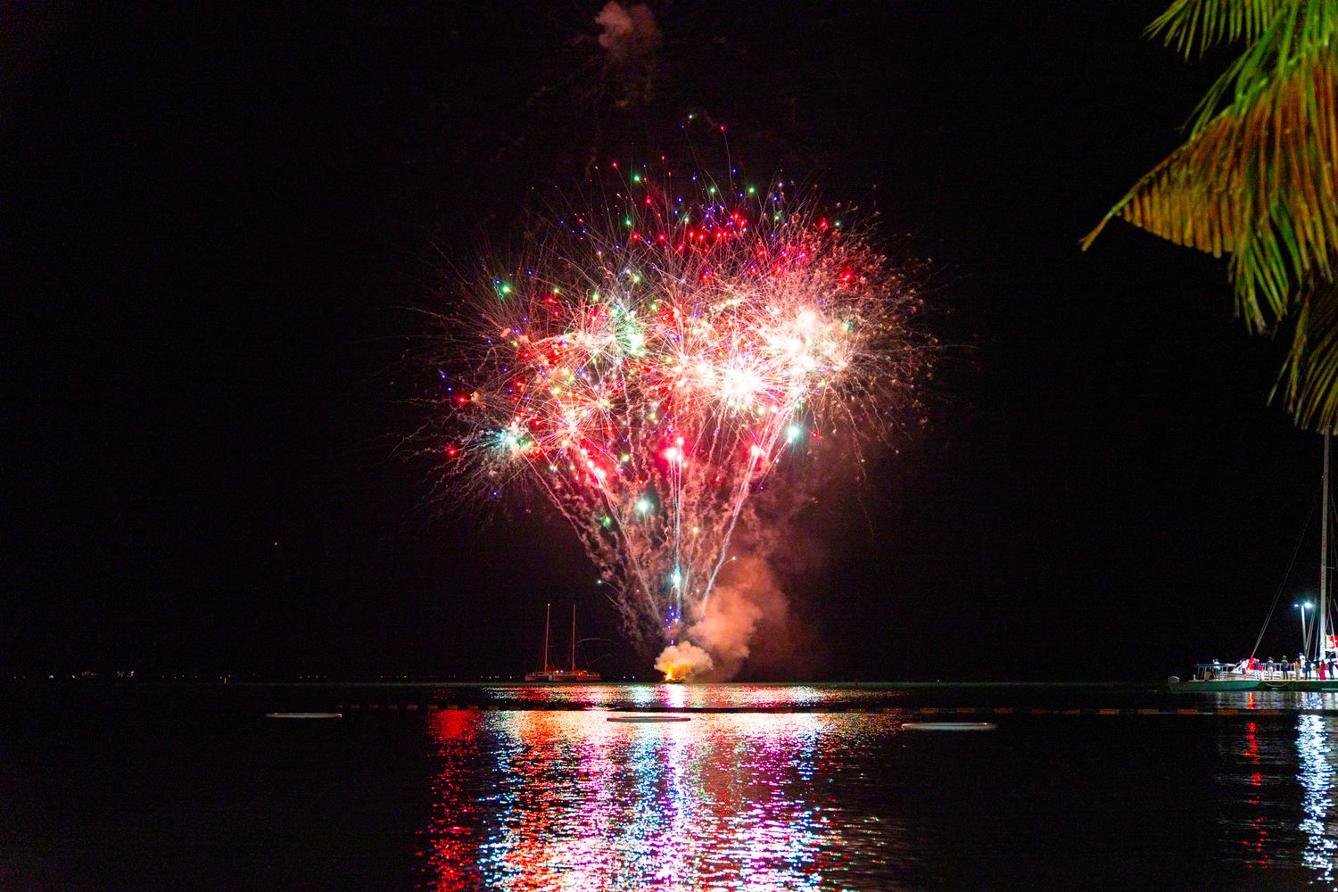 Colorful fireworks display reflecting on dark water with bright red and pink bursts in the night sky.