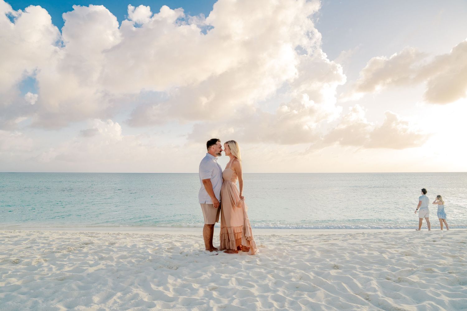 Couple standing on white sand beach at sunset with dreamy clouds and ocean view.
