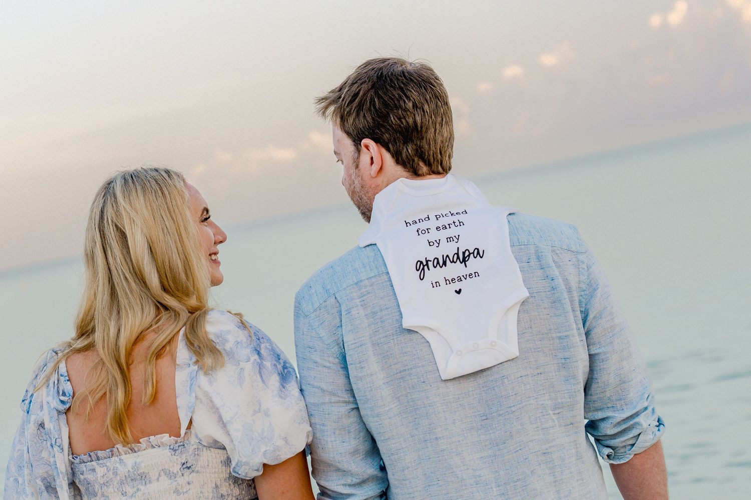 Couple in casual attire holding pregnancy announcement photos on beach at sunset.